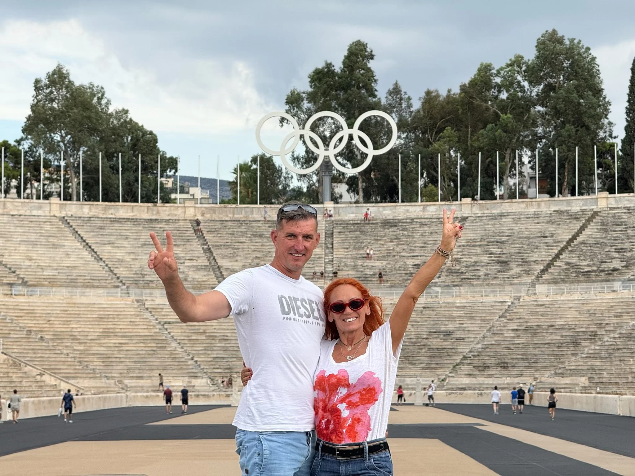 Two people posing in front of the Athens Amphitheatre with the Olympic rings set into a sign. The man is wearing sunglasses, a white T-shirt that says 'DIESEL,' and has short gray hair. The woman has red hair, wearing glasses, a white T-shirt with a pink and red artistic print, blue jeans, and a belt, making peace signs with her hands.
