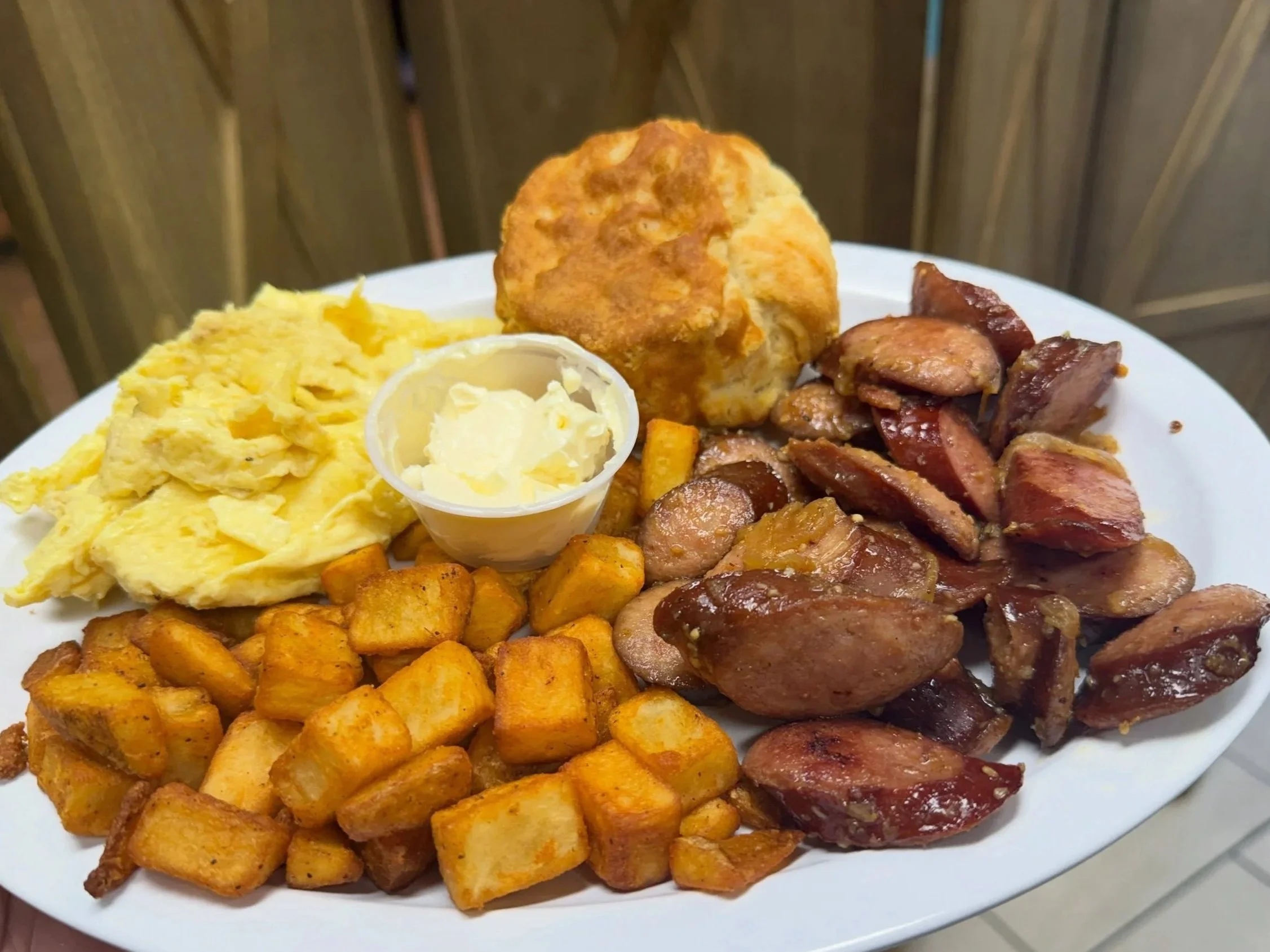 A plate of breakfast food including scrambled eggs, fried chicken, roasted sweet potatoes, biscuits with butter, and chopped sausage.