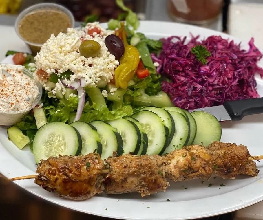 A plate with a grilled chicken skewer, cucumber slices, a mixed green salad topped with feta cheese and olives, shredded purple cabbage, and two small cups of dressing or sauces.