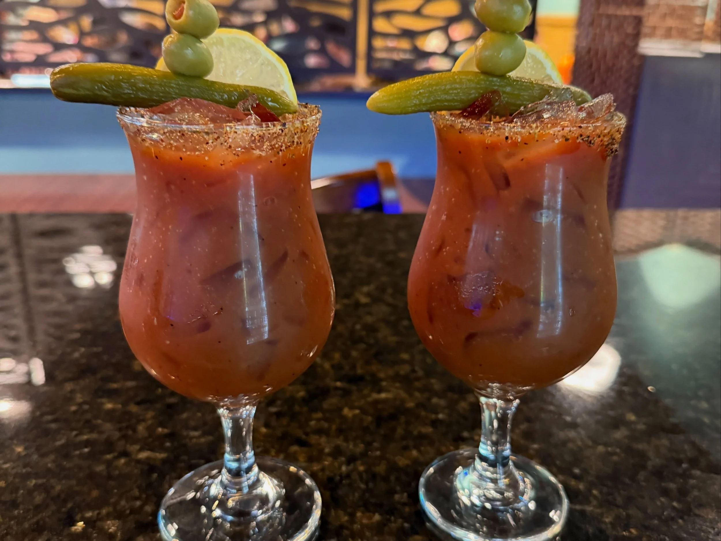 Two glasses of Mexican Michelada beer with rim salt, garnished with lemon, green chili, pickled green olives, and slices of lemon on the rim, set on a dark granite table.