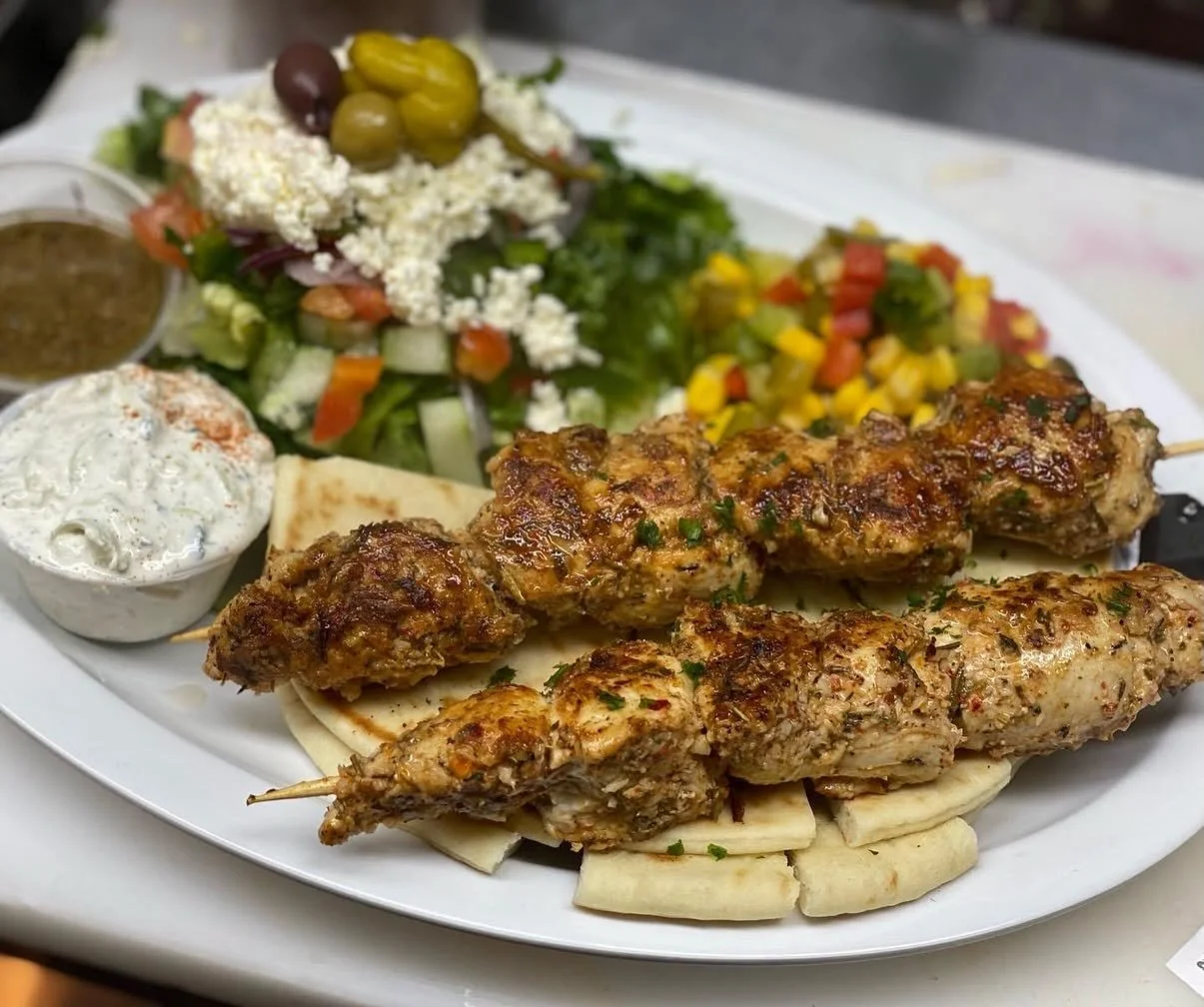 Plate with grilled chicken skewers, pita bread, salad with cucumbers, tomatoes, feta cheese, olives, and dressings, and a side of tabbouleh.