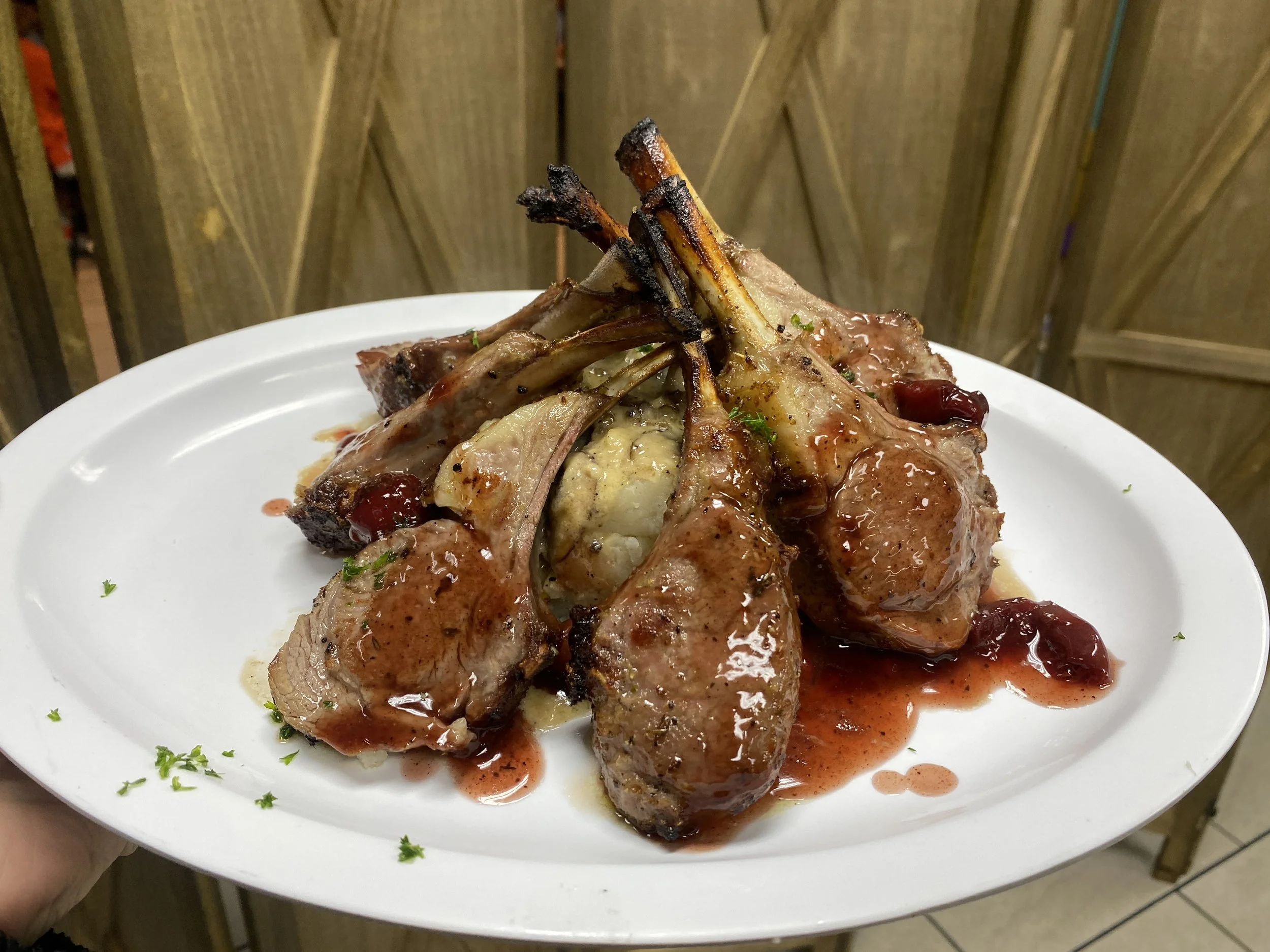 A plate with cooked lamb chops topped with a red sauce, garnished with chopped herbs, and served with a side of mashed potatoes.