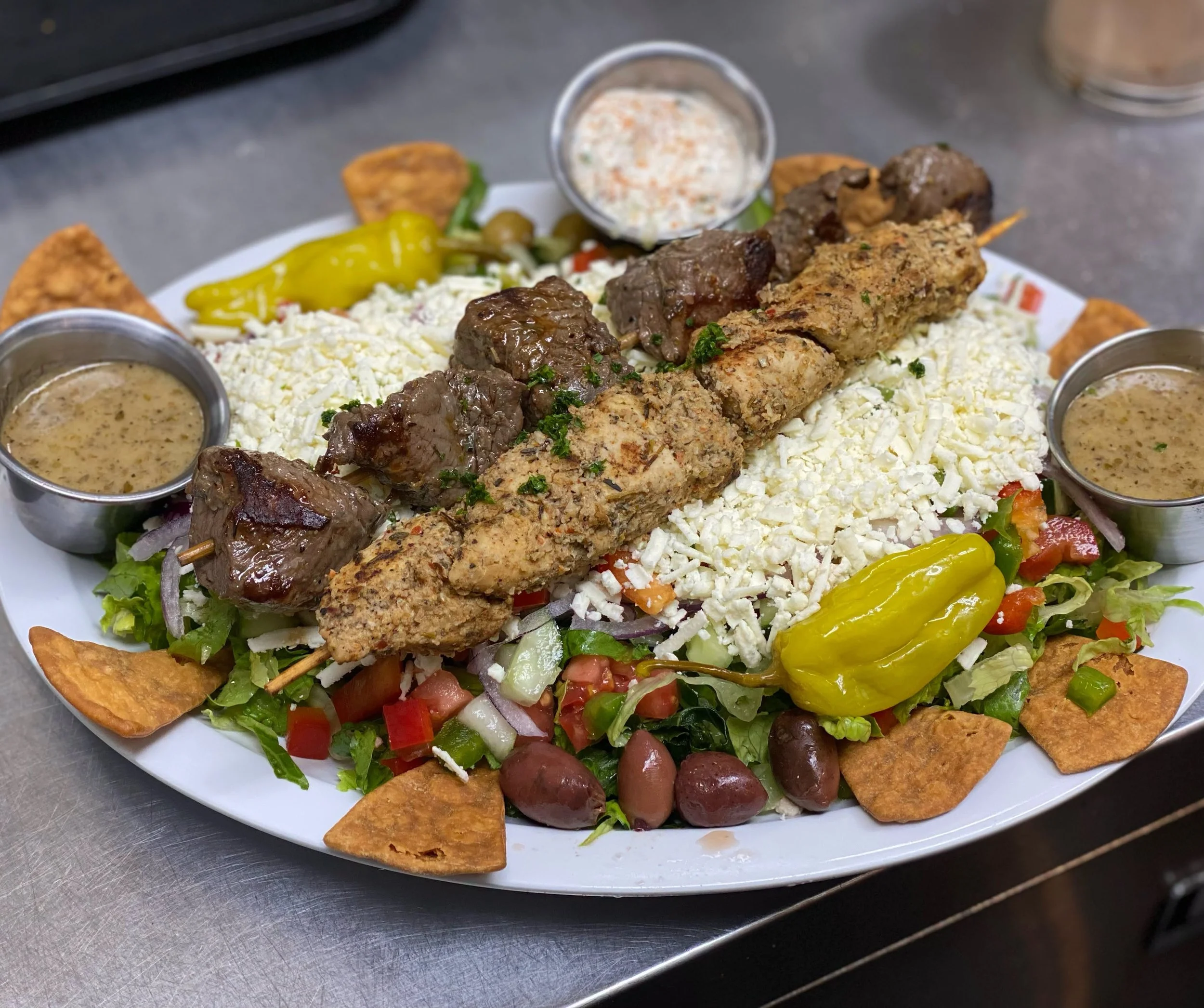 A plate of traditional Mexican food featuring grilled beef and chicken skewers, shredded cheese, lettuce, diced tomatoes, sliced jalapeños, grape tomatoes, tortilla chips, and two small containers of dipping sauce on a white oval plate.