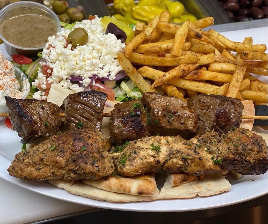 Plate of Middle Eastern food including grilled meat skewers, French fries, salad, pita bread, and a small bowl of sauce.