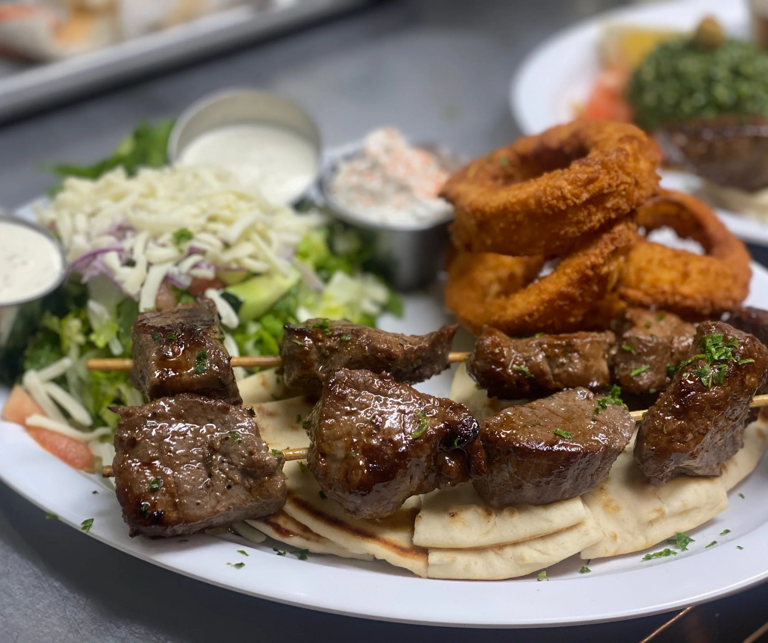 Close-up of a plate with grilled meat skewers, curly fries, shredded lettuce salad, and dipping sauces.