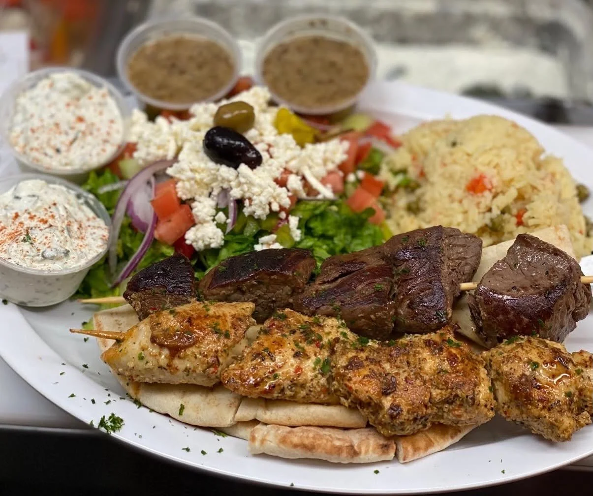 Plate of grilled meats, rice, salad with tomatoes, olives, feta cheese, and various sauces, served with pita bread.