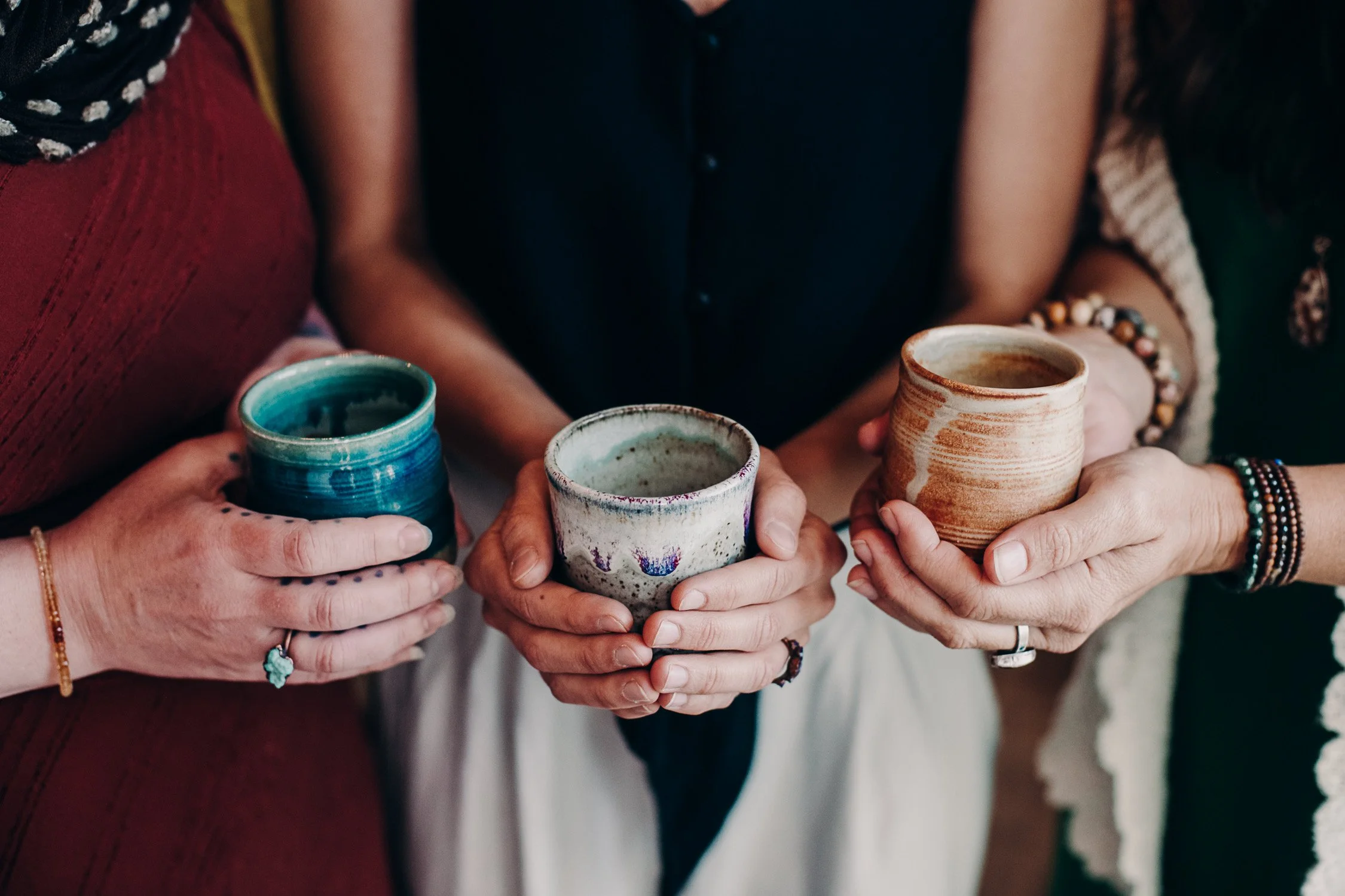 A close-up of four people holding handmade ceramic cups, showcasing different colors and designs, with some wearing bracelets and rings.