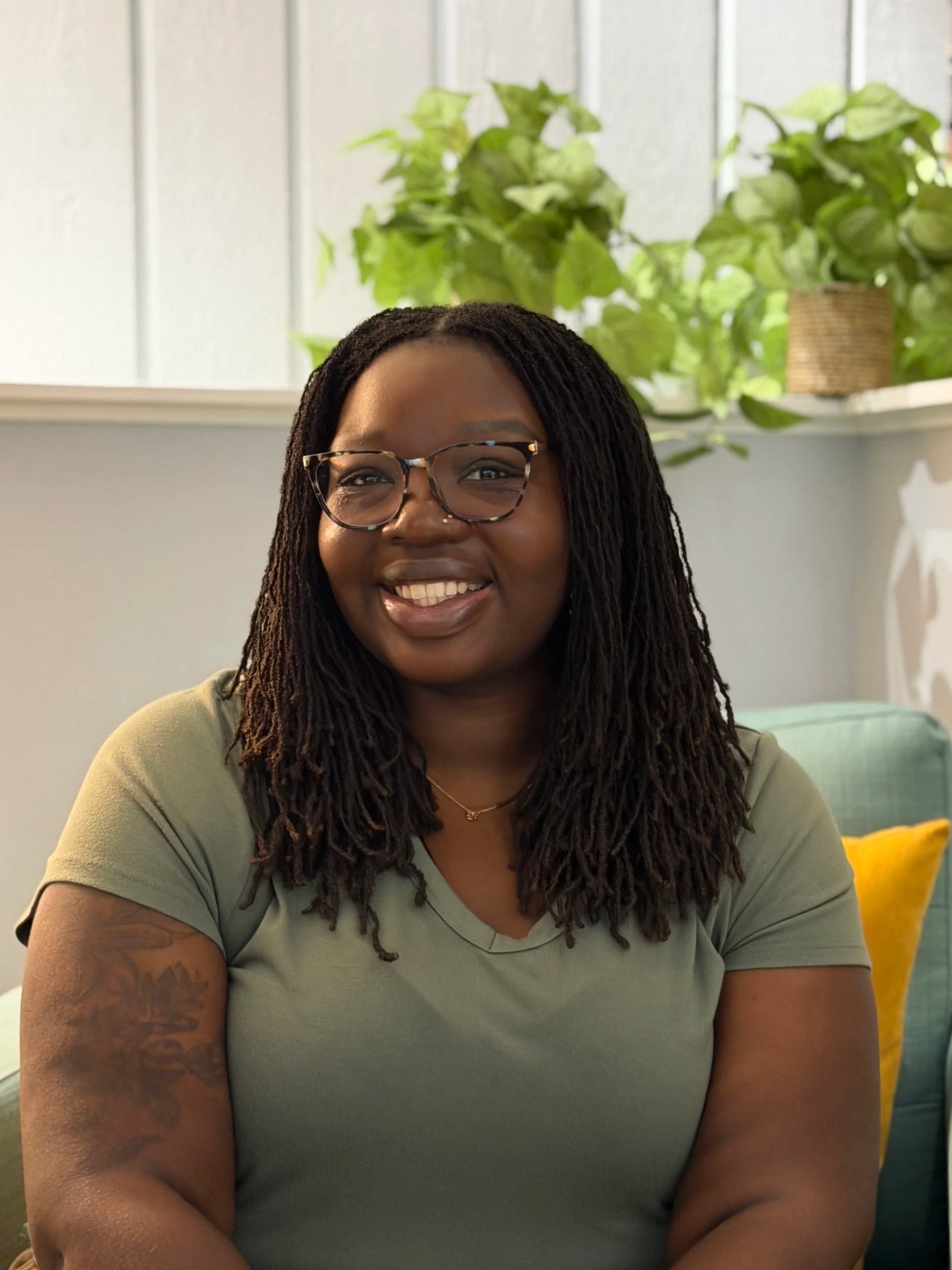 A smiling woman with long, dark, braided hair wearing glasses and a khaki shirt, sitting indoors with green plants and a window in the background.