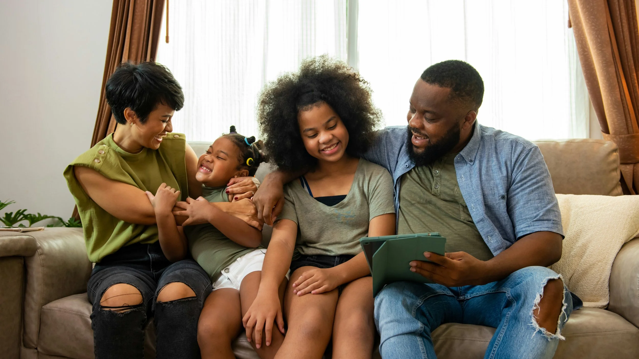 Family of five sitting on a sofa, enjoying a moment together, with a woman, two girls, and a man looking at a tablet.
