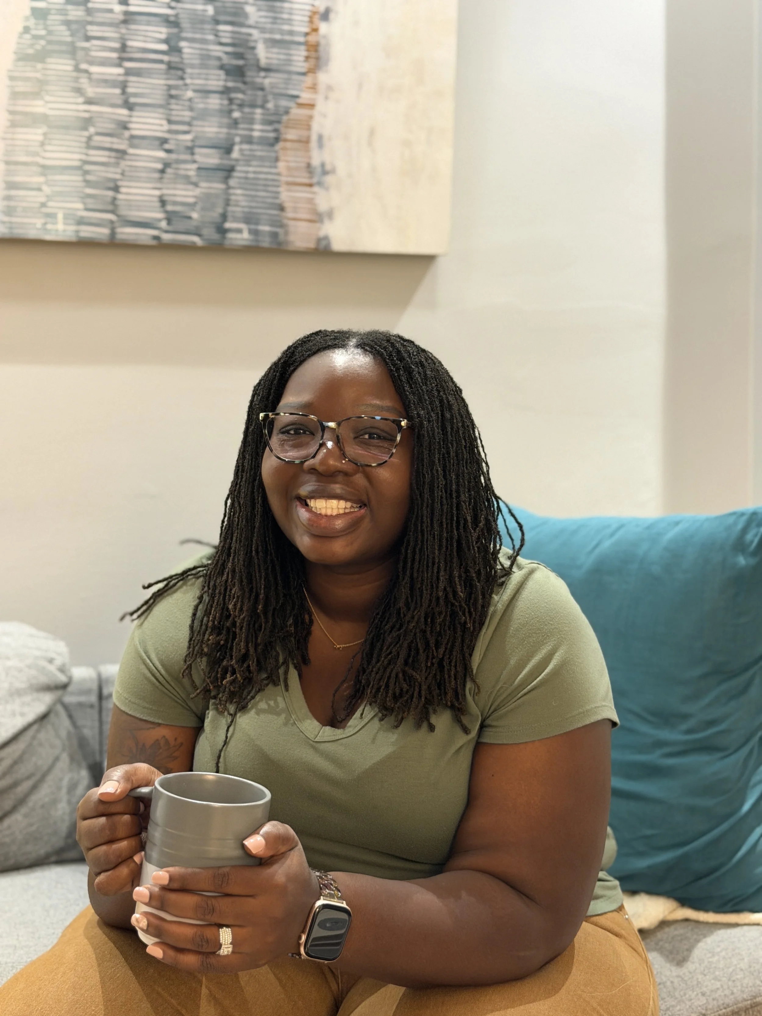 A woman with long dreadlocks, glasses, and a wide smile, sitting on the floor inside a home, holding a gray mug with both hands. She is wearing a light green shirt, brown pants, a watch, and a ring, with a beige couch and a large blue cushion behind her.