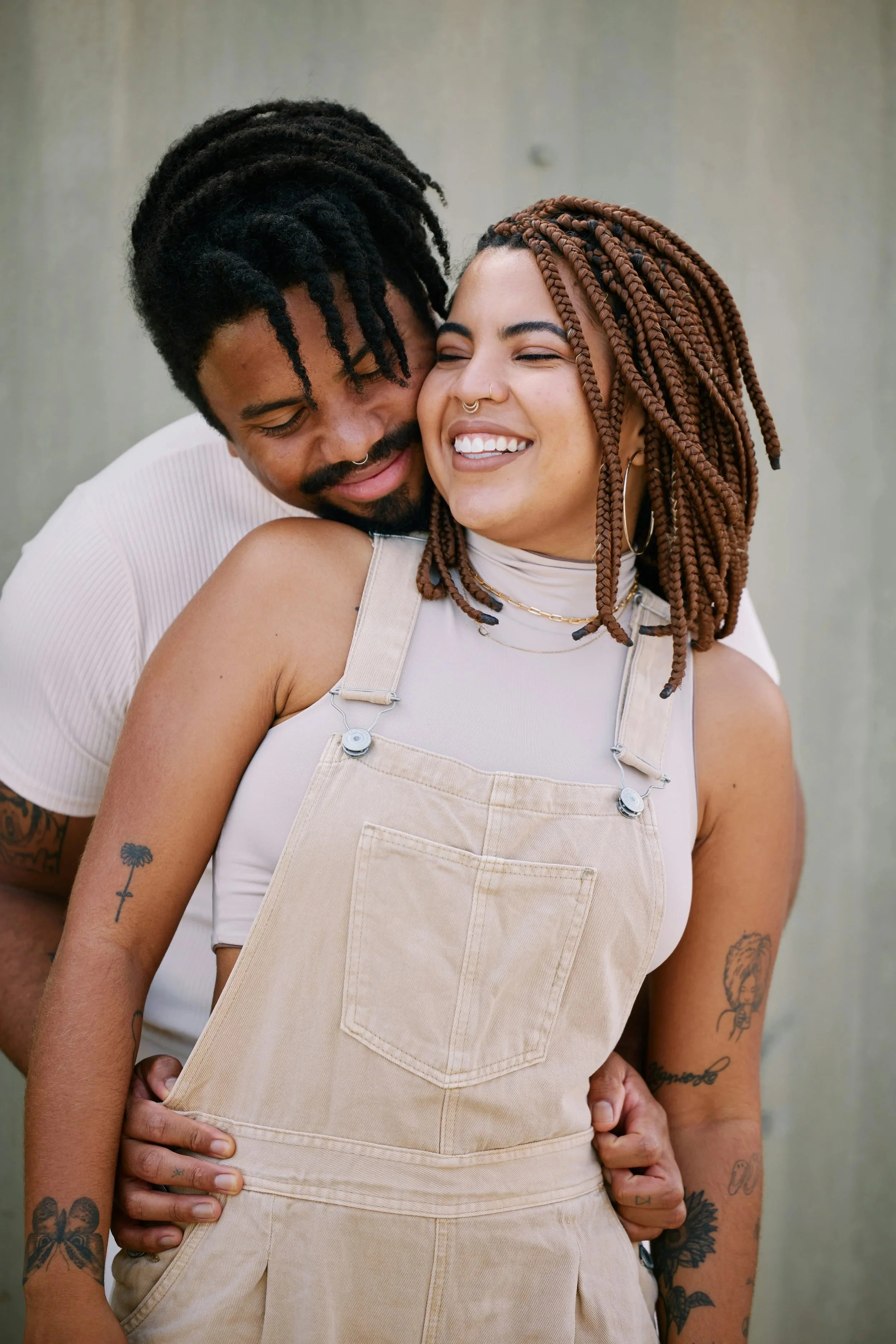 A happy couple hugging and smiling together outside, with the woman in beige overalls and braided hair, and the man with dreadlocks and a septum piercing.
