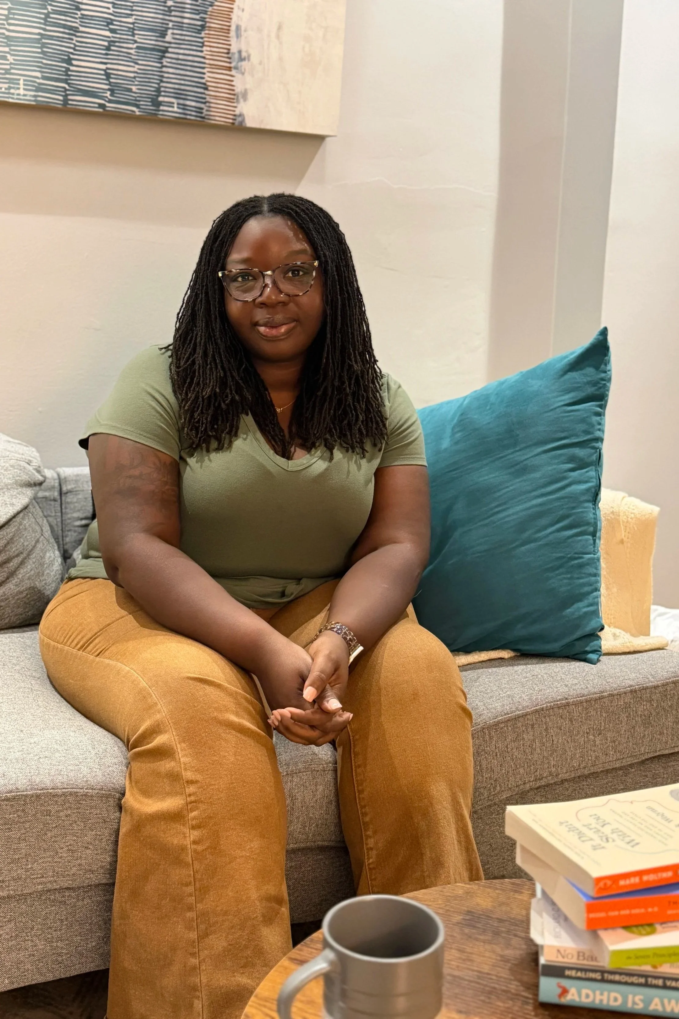 A woman with glasses and long dreadlocks sitting on a couch in a living room with beige walls, with a blue pillow behind her, wearing an olive green shirt and mustard-colored pants. There is a pile of books and two coffee mugs on a wooden table in front of her.