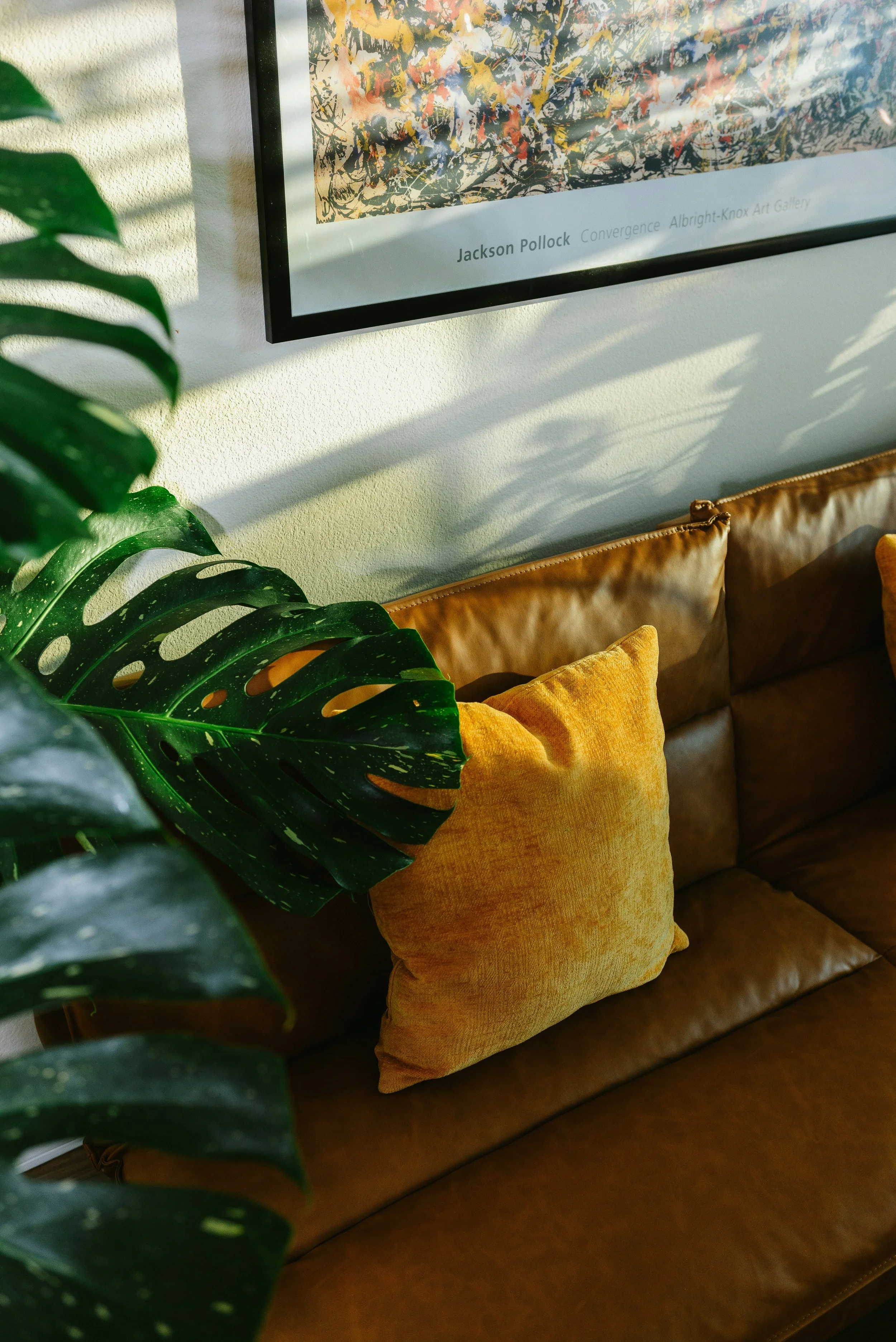A framed abstract artwork hangs on a wall above a brown leather couch adorned with mustard-colored pillows. A large green monstera plant with distinctive split leaves partially obscures the view of the couch and frame.