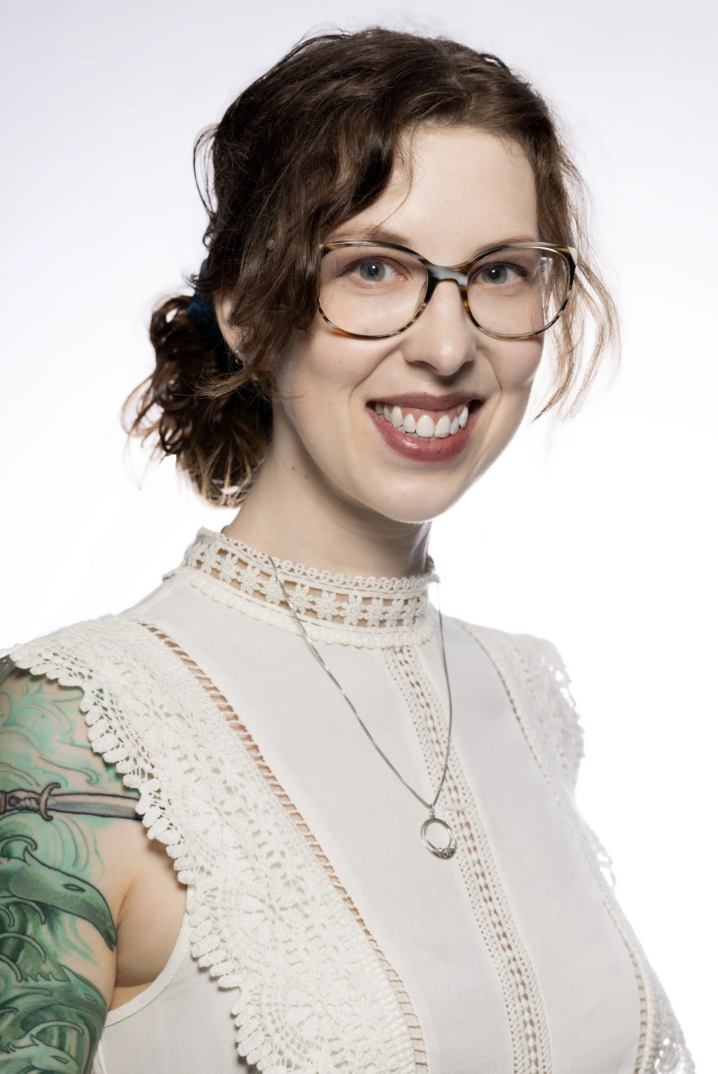 A smiling woman with curly brown hair, wearing glasses, a white lace top, a necklace with a circular pendant, and a colorful tattoo on her arm, against a plain white background.