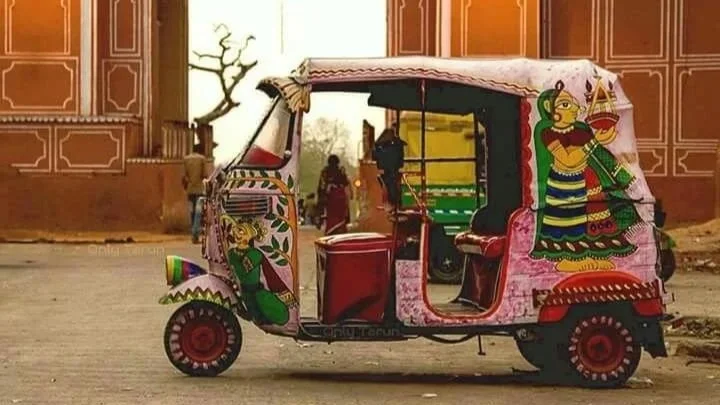 Colorfully decorated auto rickshaw with artistic designs and figures, parked in front of a historical gate with a tree in the background.