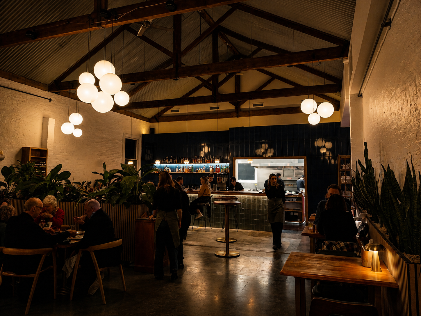 A cozy restaurant interior with white brick walls, wooden beams, hanging spherical lights, and a bar area with bar stools. Several diners are sitting at tables, and waitstaff are serving. The overall ambiance is warm and inviting.
