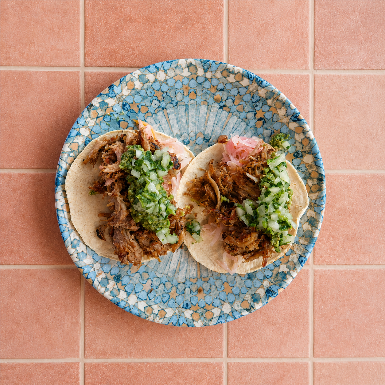 Two tacos on a decorative blue plate, filled with shredded meat, chopped green onions, and shredded pink pickled vegetables, set on a pink tiled surface.