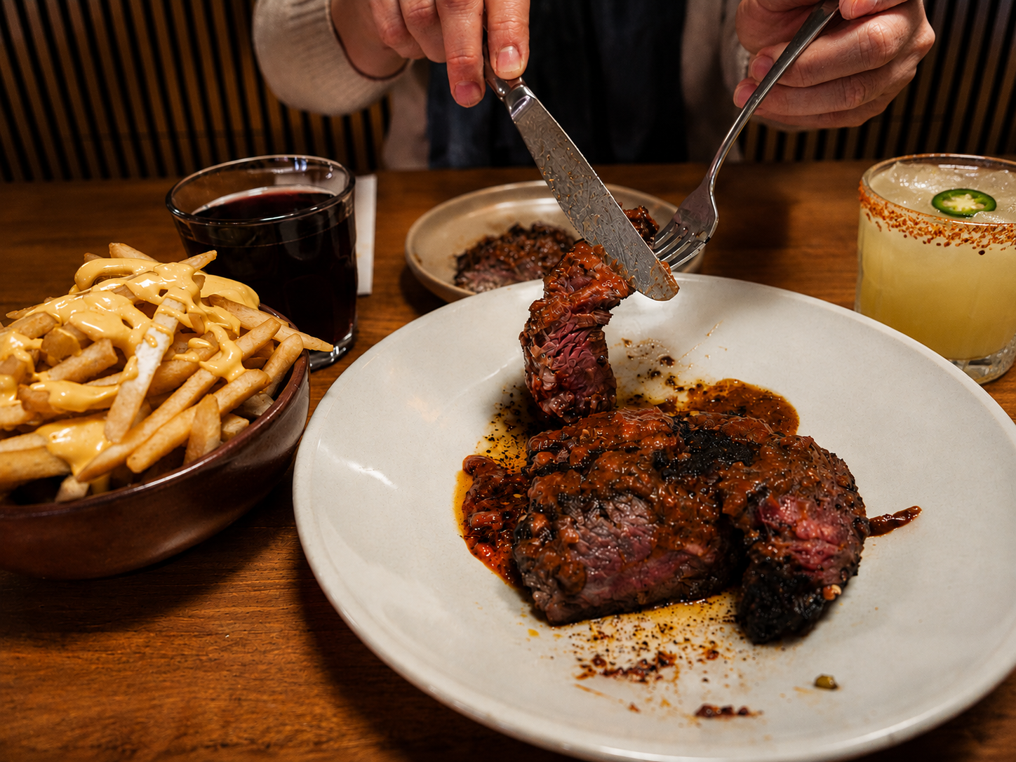 Person cutting into a cooked, sauced steak with French fries topped with cheese, a glass of red wine, a yellow cocktail with jalapeño garnish, and a small dish of sauce on a wooden table.