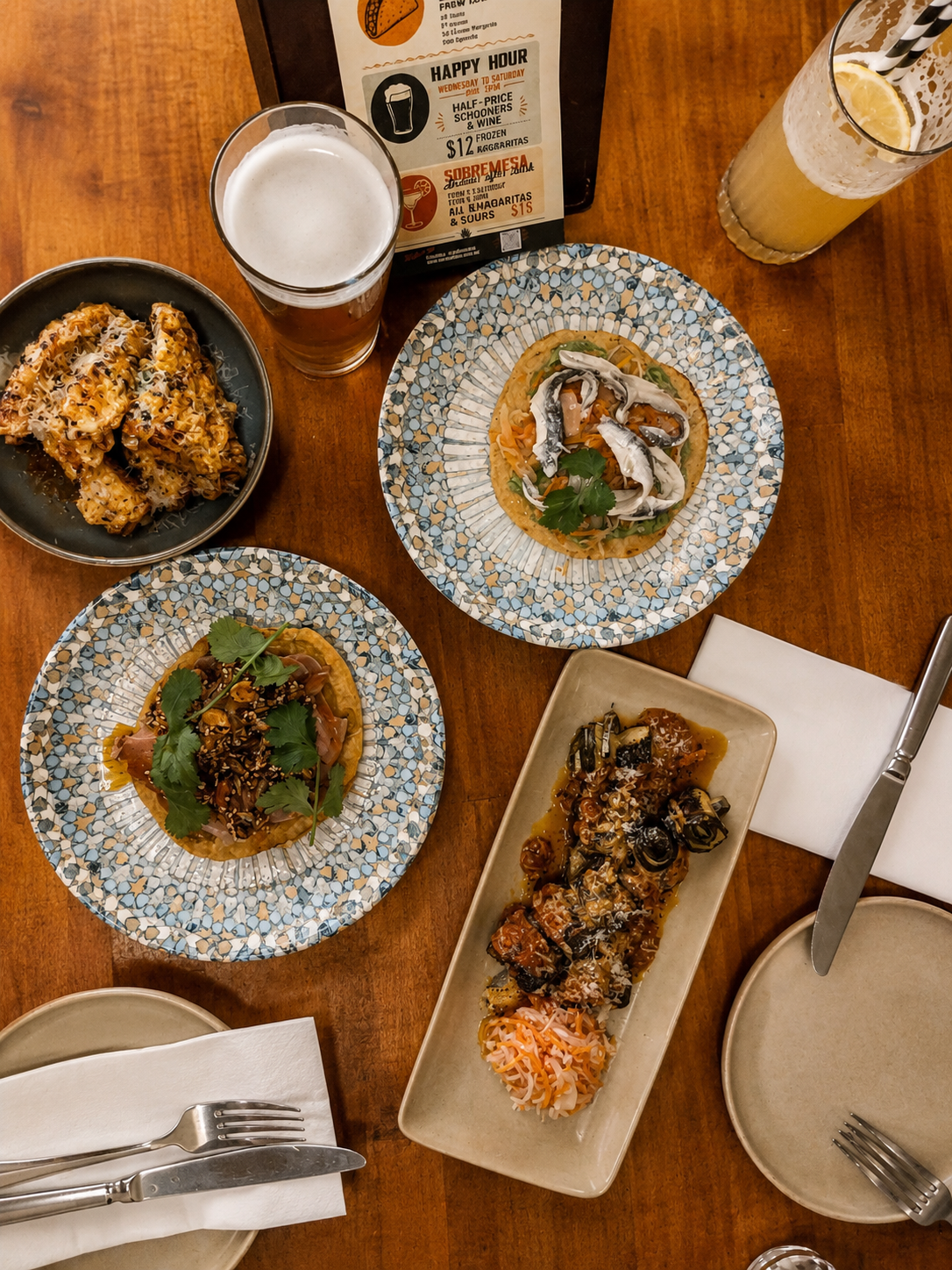 A table with various Latin Americandishes, including anchovies tortilla topped with cilantro, corn ribs, a plate of tuna belly with sesame seeds and peanuts, and two glasses of beverages, one with lemon slices, on a wooden table.