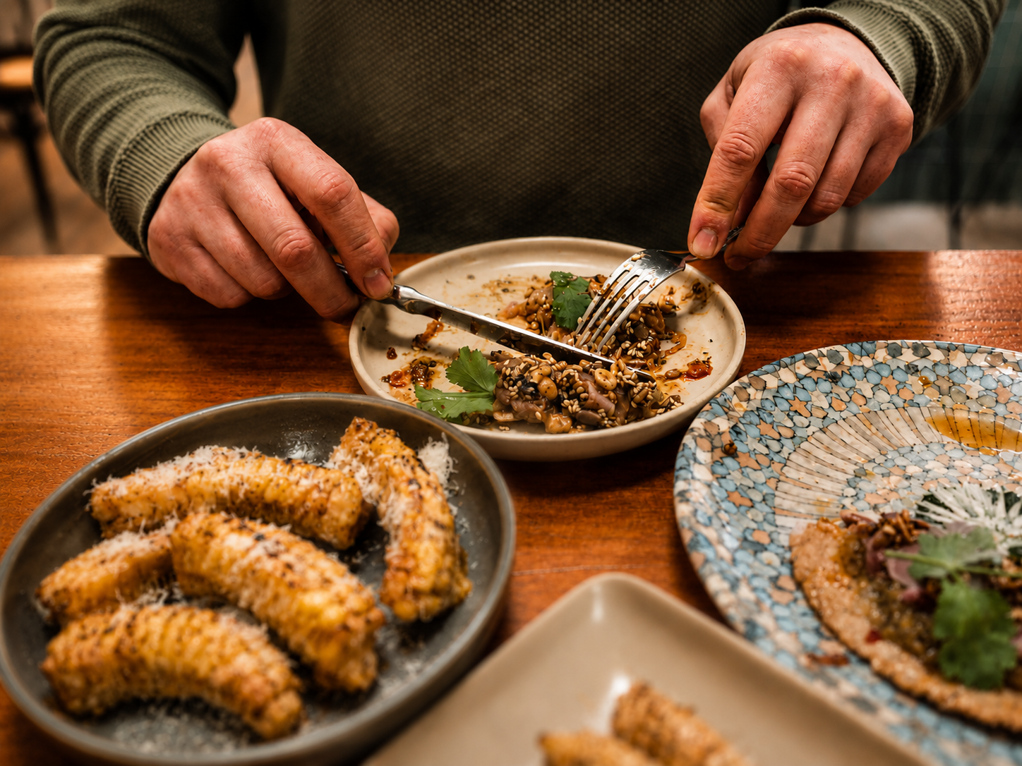 Hands holding a fork and spoon eating a dish of tuna belly with salsa macha on a crispy homemade tortilla with cilantro, with plates of fried food and other dishes on a wooden table.