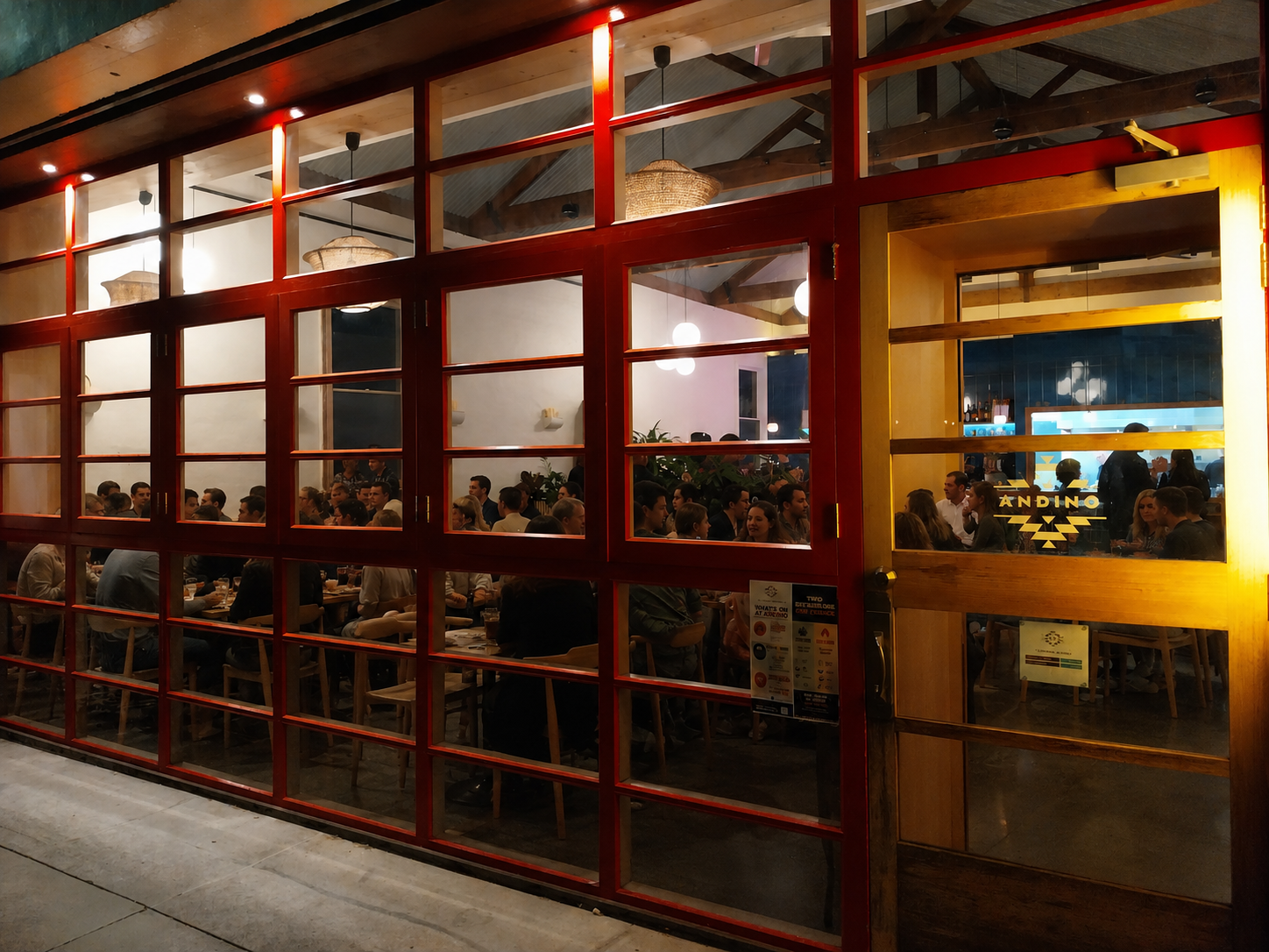 View through large red-framed glass window of a busy restaurant interior filled with seated customers, warm lighting, and wooden decor.