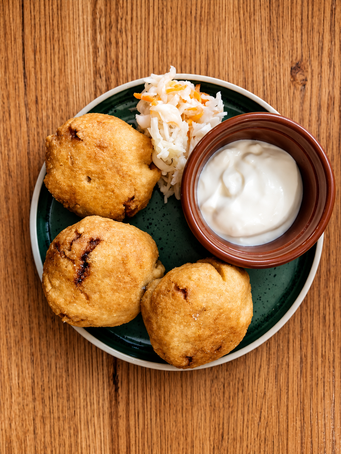Plate with three fried cauliflower pupusass, pickled slaw and carrots, and a cup of white creamy garlic dip, on a wooden table.