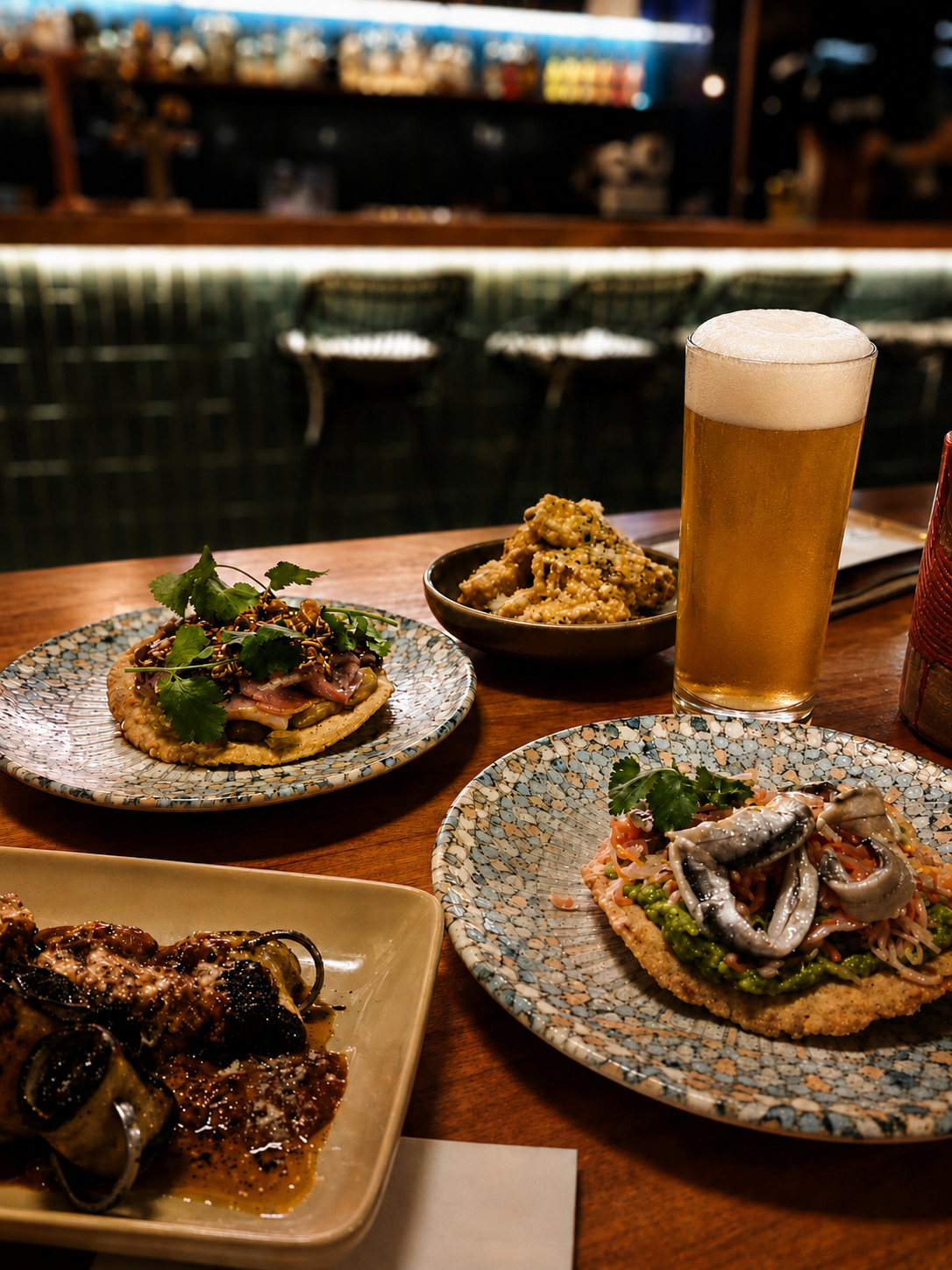 A table with various Latin American dishes, like Anchovies tostada, corn rib, meat skewers, and tuna crudo. There's also a beer in a tall glass, on a wooden table in a restaurant with a dark background.