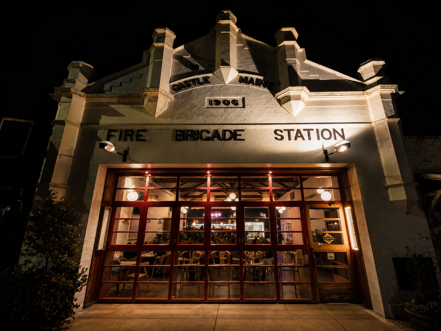 Night view of Bar Andino in the historic Castlemaine Fire Brigade Station built in 1906, with illuminated signage and large glass doors showing the interior with chairs and hanging lights.