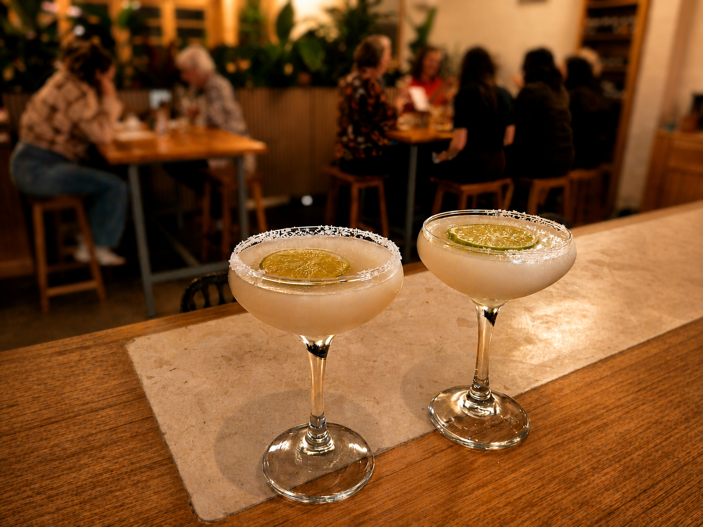 Two frozen margarita cocktails with salted rims and lime slices on a wooden bar counter in a restaurant, with people sitting at tables in the background.