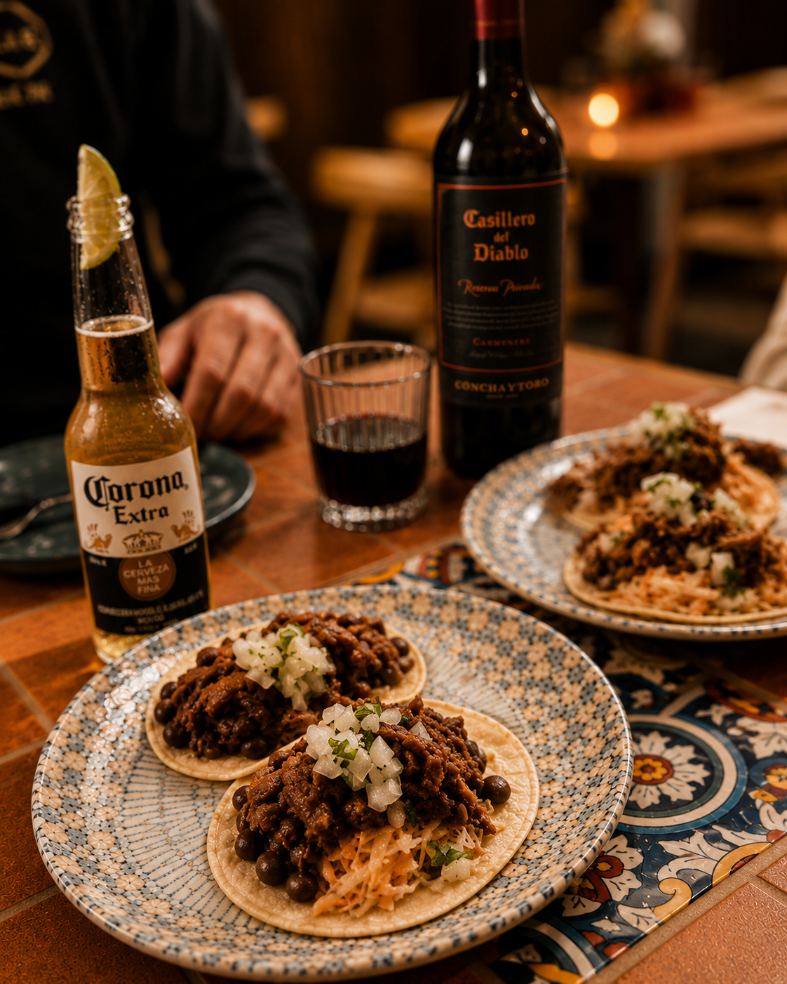 Two plates of tacos with pulled pork, beans, and chopped onions on a decorative ceramic plate. A bottle of red wine, a glass of red wine, and a bottle of Corona Extra beer are on the wooden table. Part of a person’s arm and hand are visible.