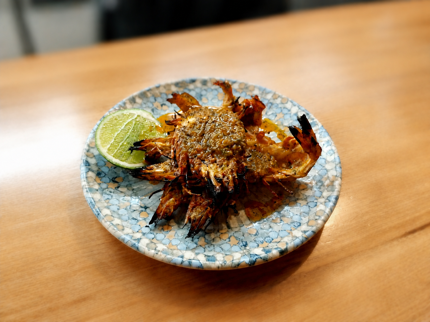 Grilled whole prawns topped with sauce and chopped garlic, served on a decorative plate with lime wedge, on a wooden table.