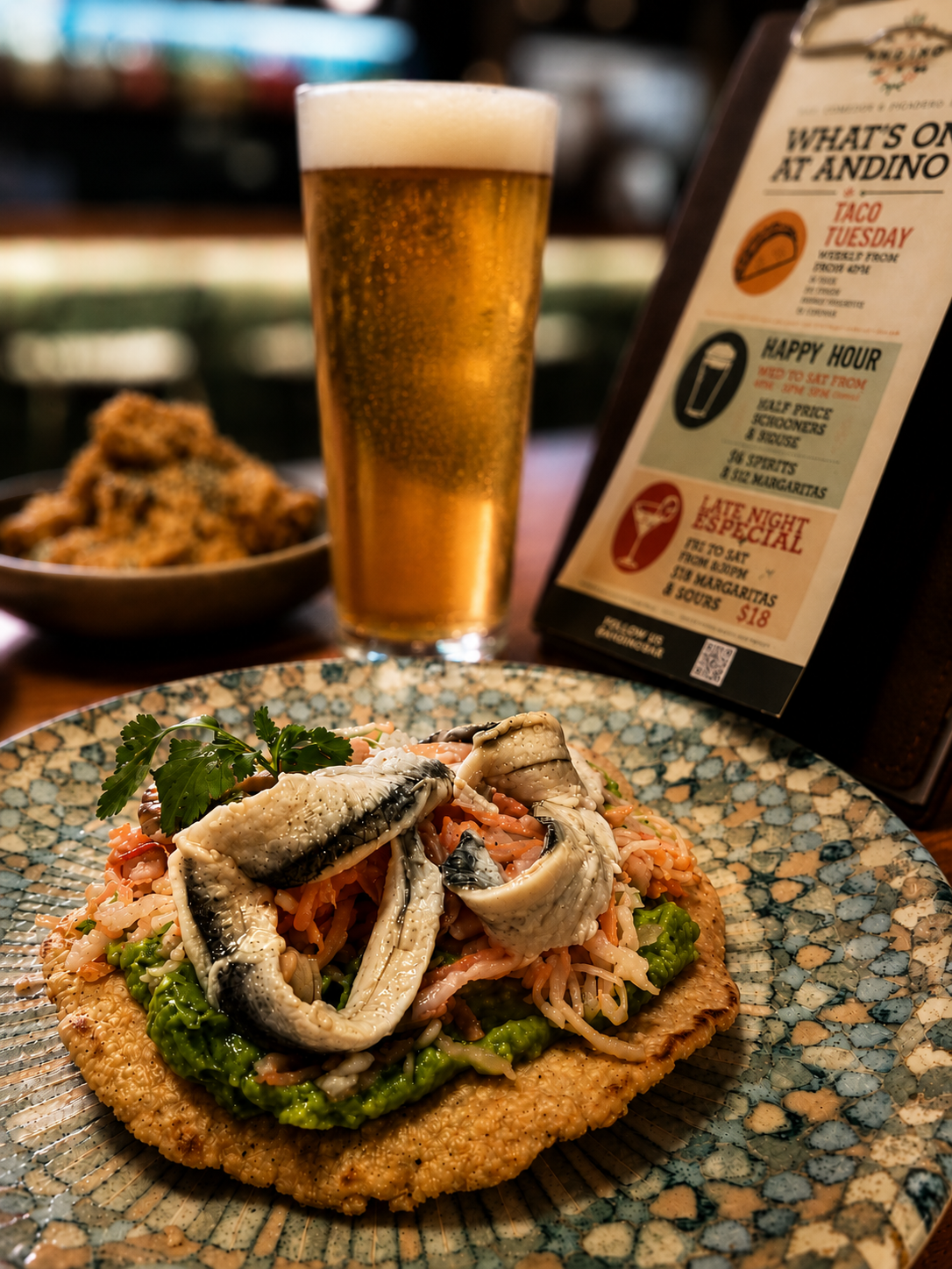 A plate of fried white anchovies with shredded vegetables and caviar garnished with parsley, accompanied by a glass of beer, with a bowl of grilled corn and a menu in the background at a restaurant.