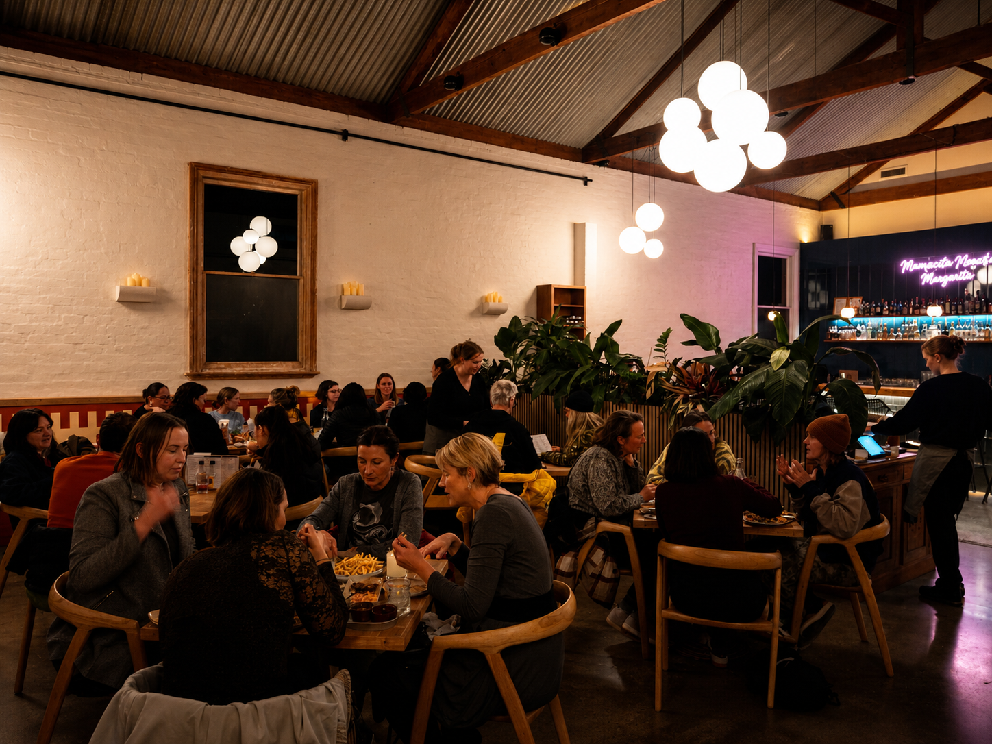 A lively restaurant scene with patrons dining, chatting, and waitstaff serving. Decor includes white brick walls, large light fixtures, and lush greenery. A bar area on the right features neon lighting and bottles.