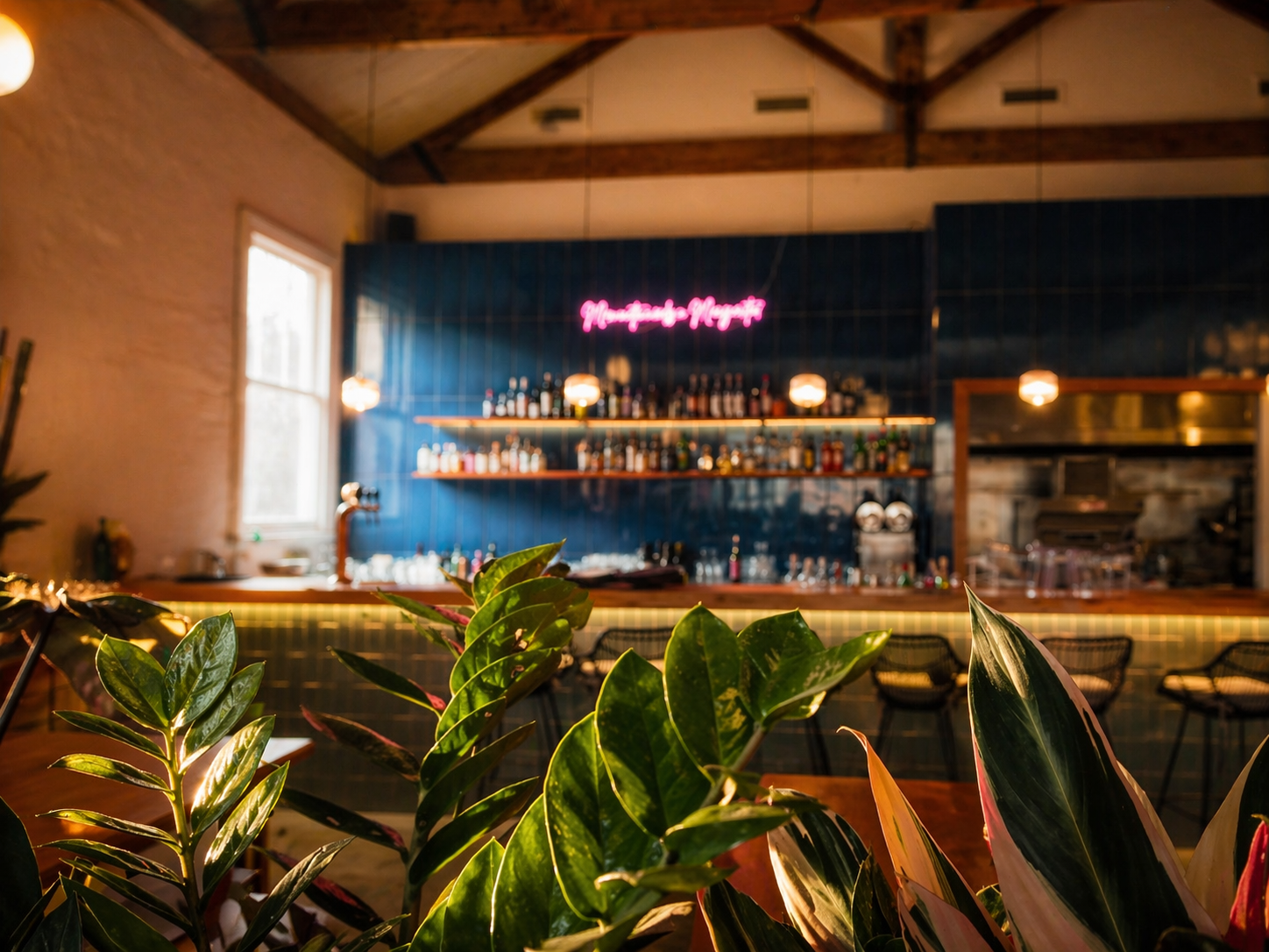 Indoor bar with a neon pink sign, shelves with bottles behind the bar, and plants in the foreground.