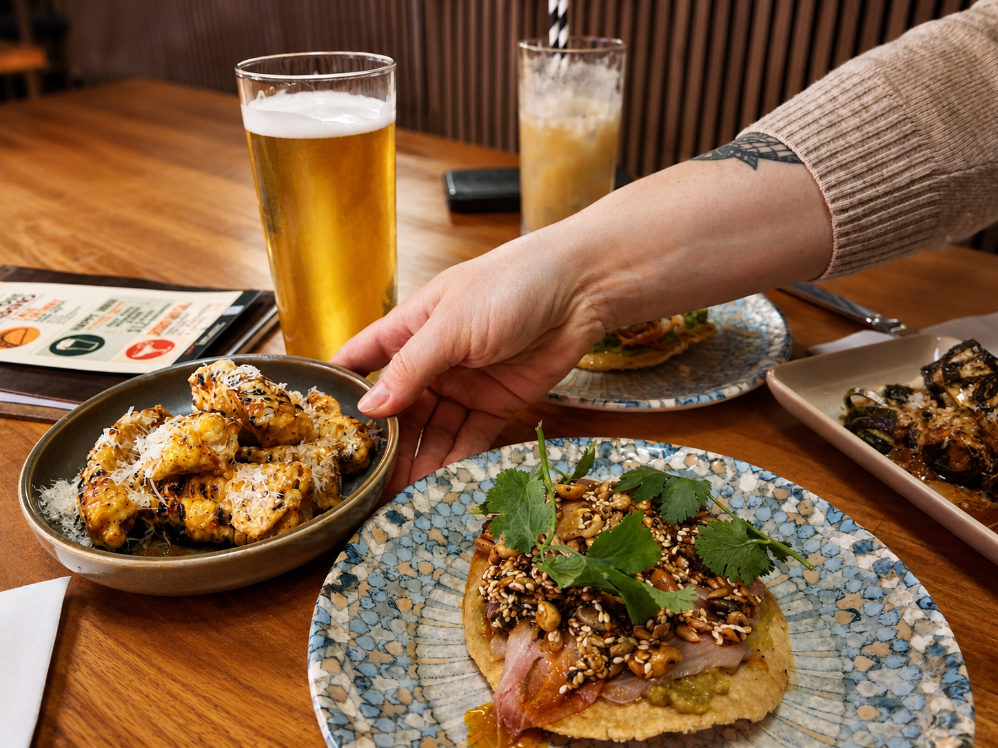 Person reaching for a bowl of corn ribs, and a plate of tuna crudo with sesame seeds and shredded cheese, around glasses of beer on a wooden table.