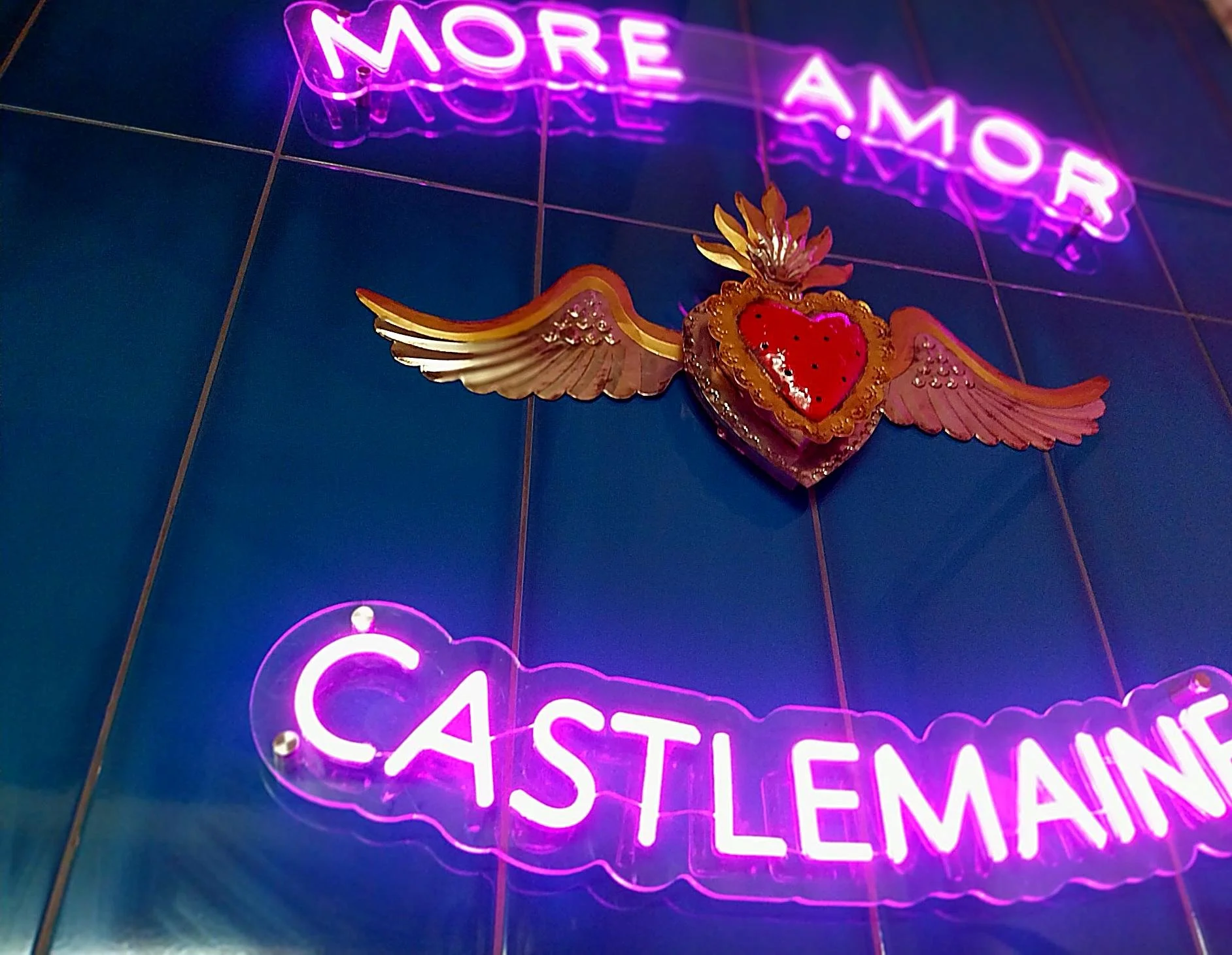 Neon sign in pink and purple lights saying 'MORE AMOR'  and 'CASTLEMAINE' on a wall with dark blue tiles, with a decorative heart with a flaming crown and wings below the sign.