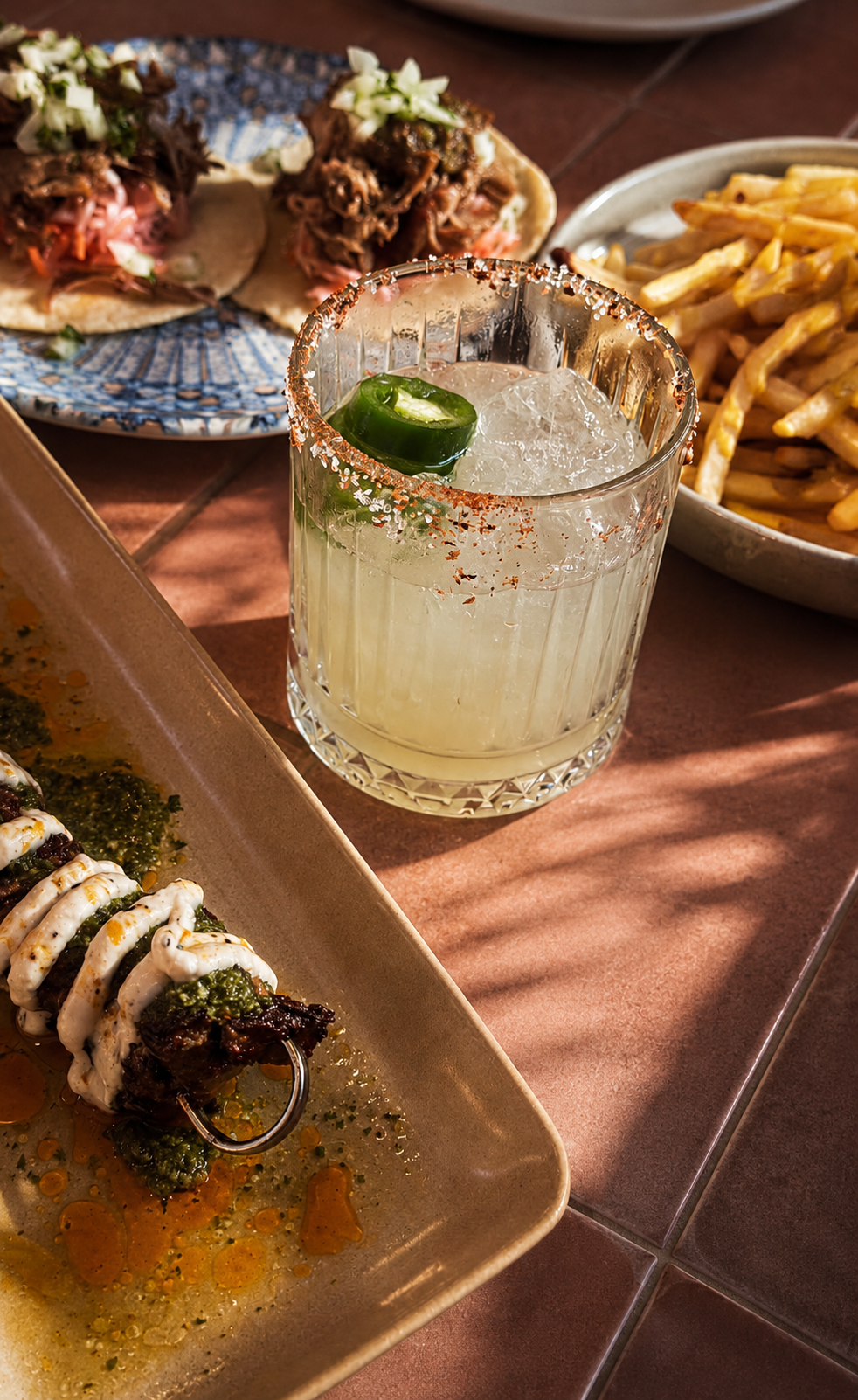 A glass of margarita with a lime wedge and salt rim, a plate of tacos with beef and toppings, a bowl of French fries, and a platter of grilled meat with sauce on a pinkish-brown table.