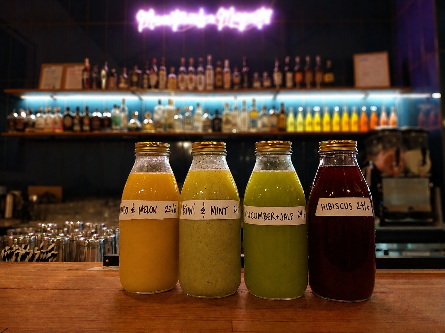 Four bottles of fresh juice with different flavors and handwritten labels, placed on a wooden bar counter in front of a liquor shelf in a dimly lit bar or cafe setting.
