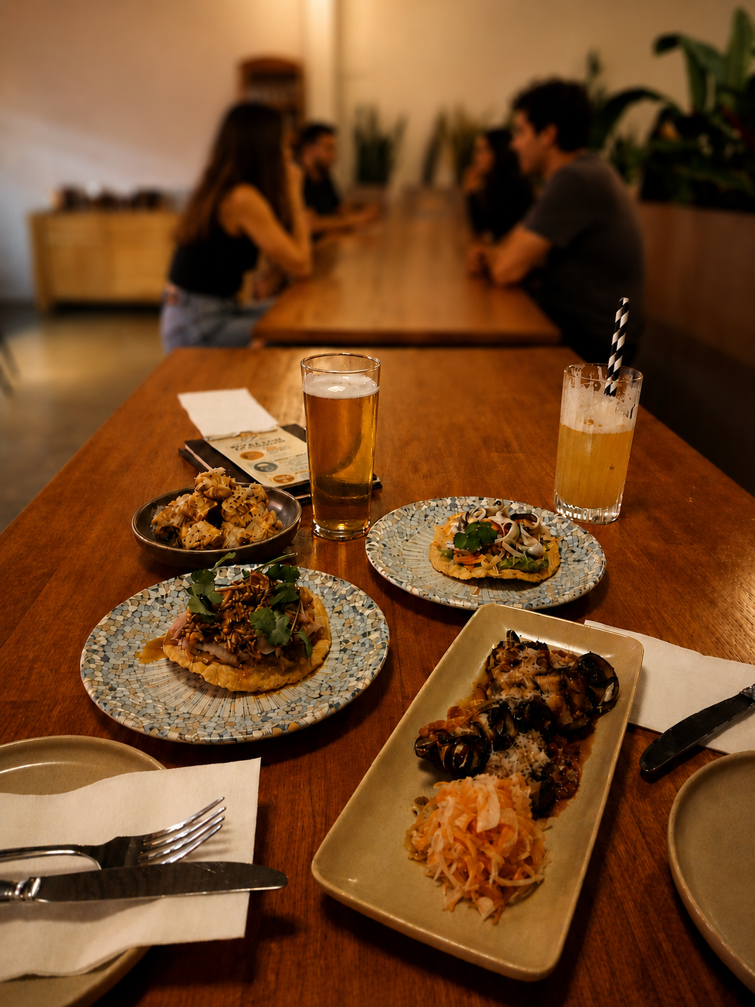A dinner table with three plates, tuna crudo, anchovies tostada, and ,eat skewer, two drinks, and utensils, with blurred people sitting in the background.