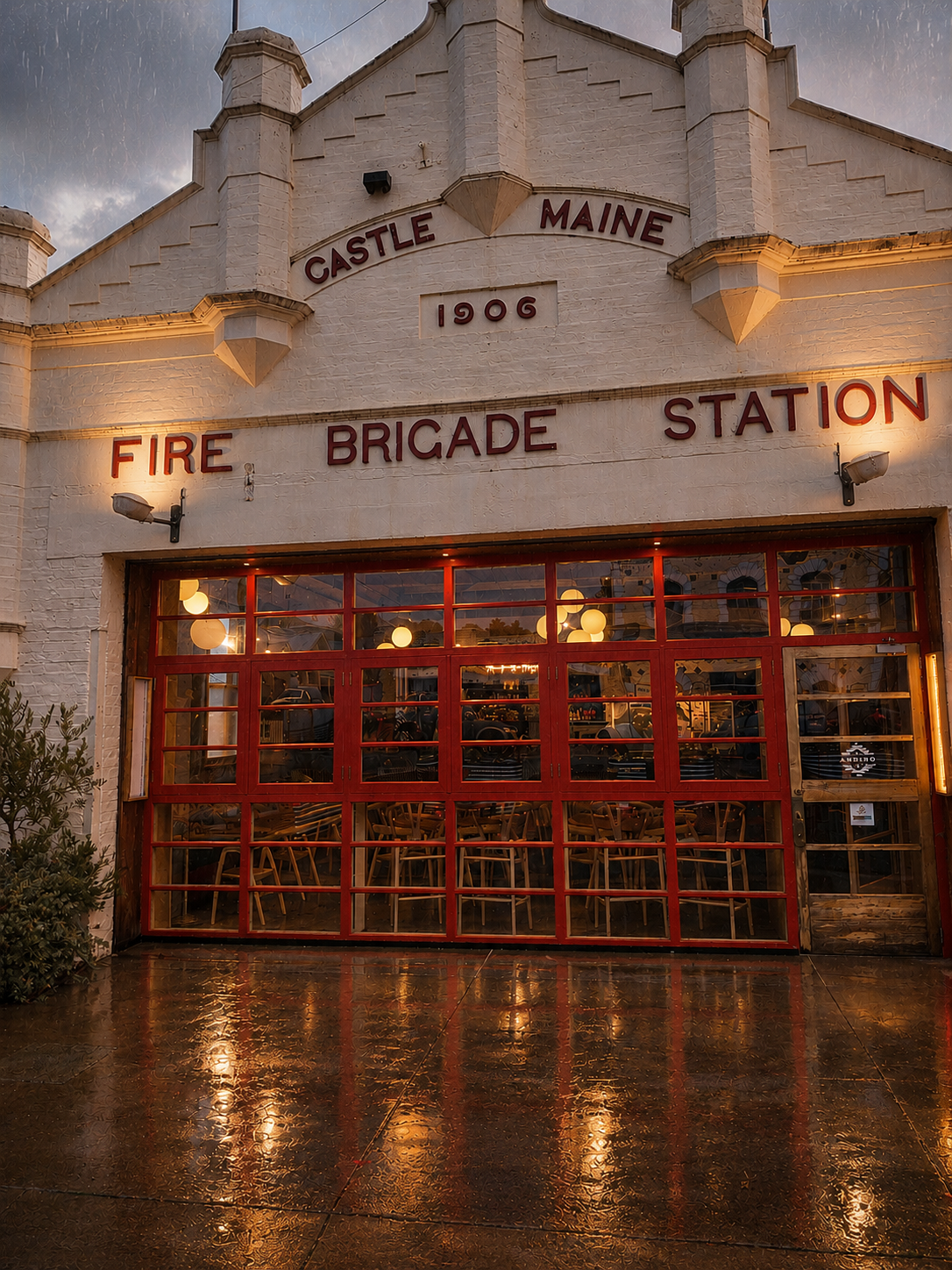 The exterior of a historic fire station built in 1906, home of Bar Andino, with a large glass garage door, red framing, and signage reading 'Castle Maine 1906 Fire Brigade Station' on the facade, illuminated by exterior lights during evening or dusk.