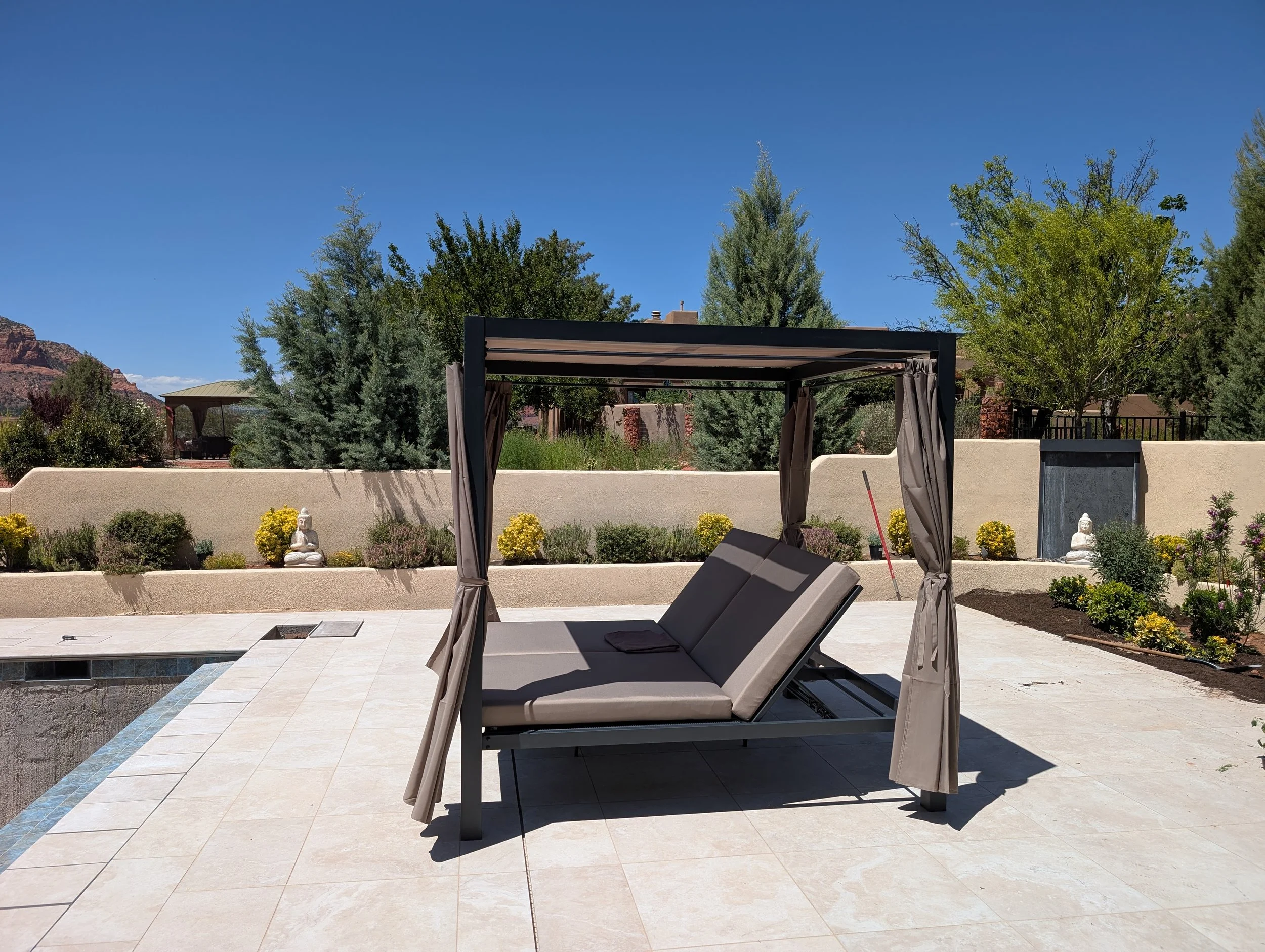 A backyard patio featuring a canopy bed with curtains, lounge chairs, a built-in pool or hot tub, decorative Buddha statues, and landscaped garden with bushes and trees, under a clear blue sky.