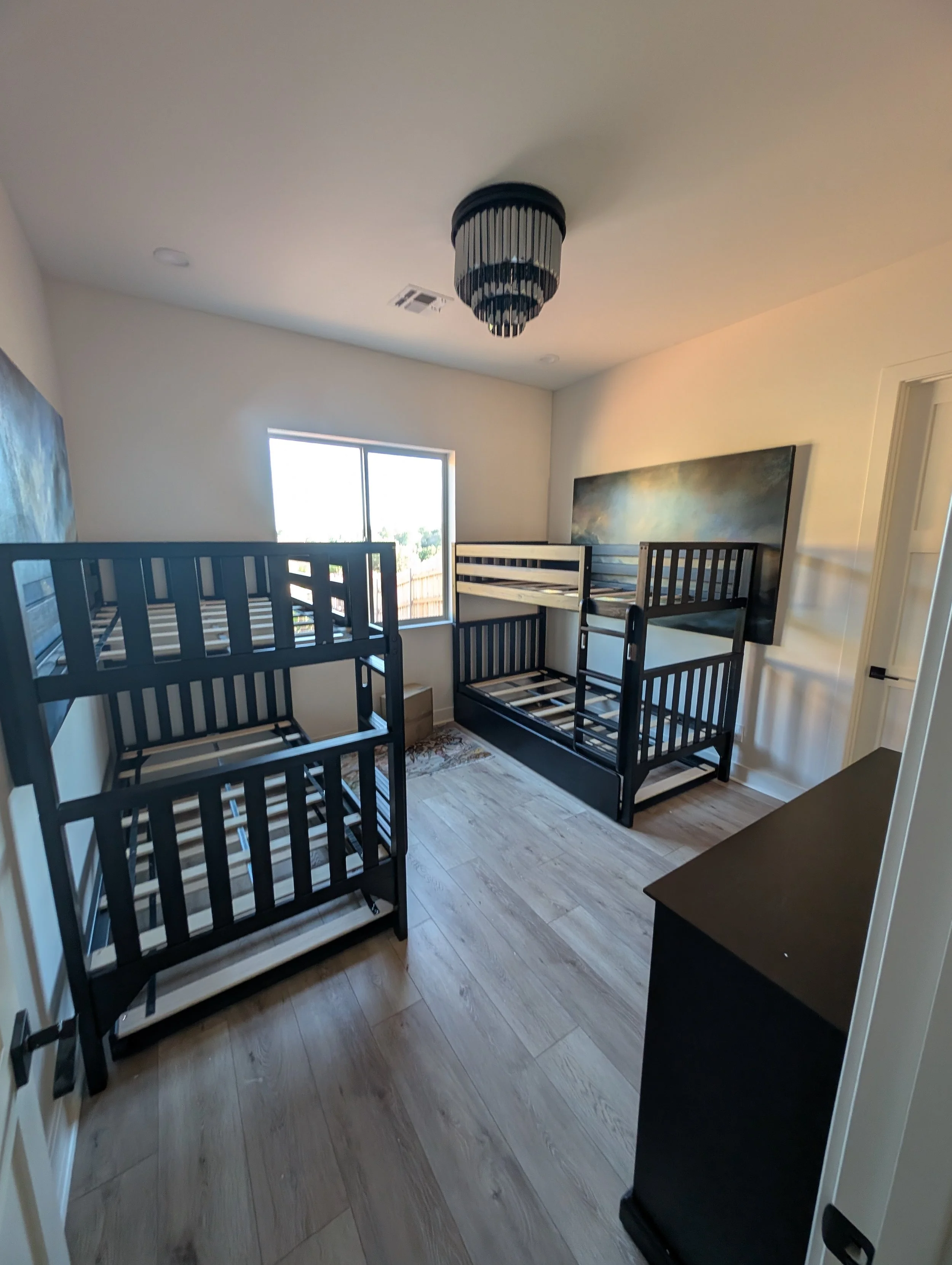 A room with three identical black bunk beds, a large window, a colorful abstract painting on the wall, and light-colored wooden flooring.