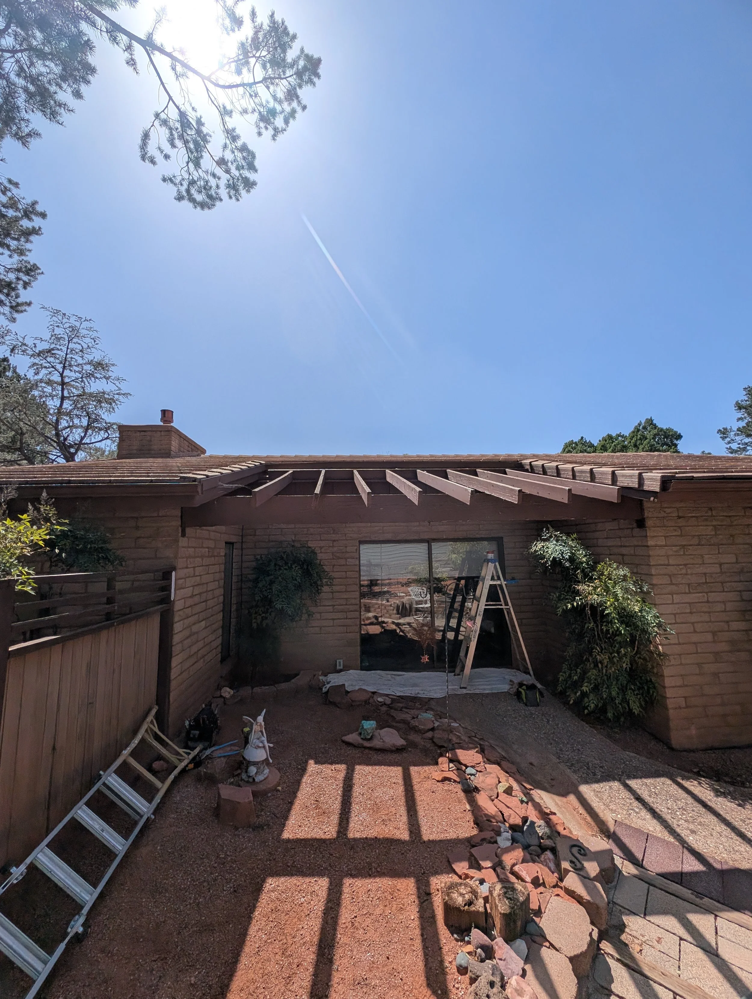 Backyard scene with a house, roof shingles, a tall ladder leaning against the house, open space for yard work, some gardening supplies, rocks, and shadows cast by a pergola or patio cover. Trees are visible in the background under clear blue sky.