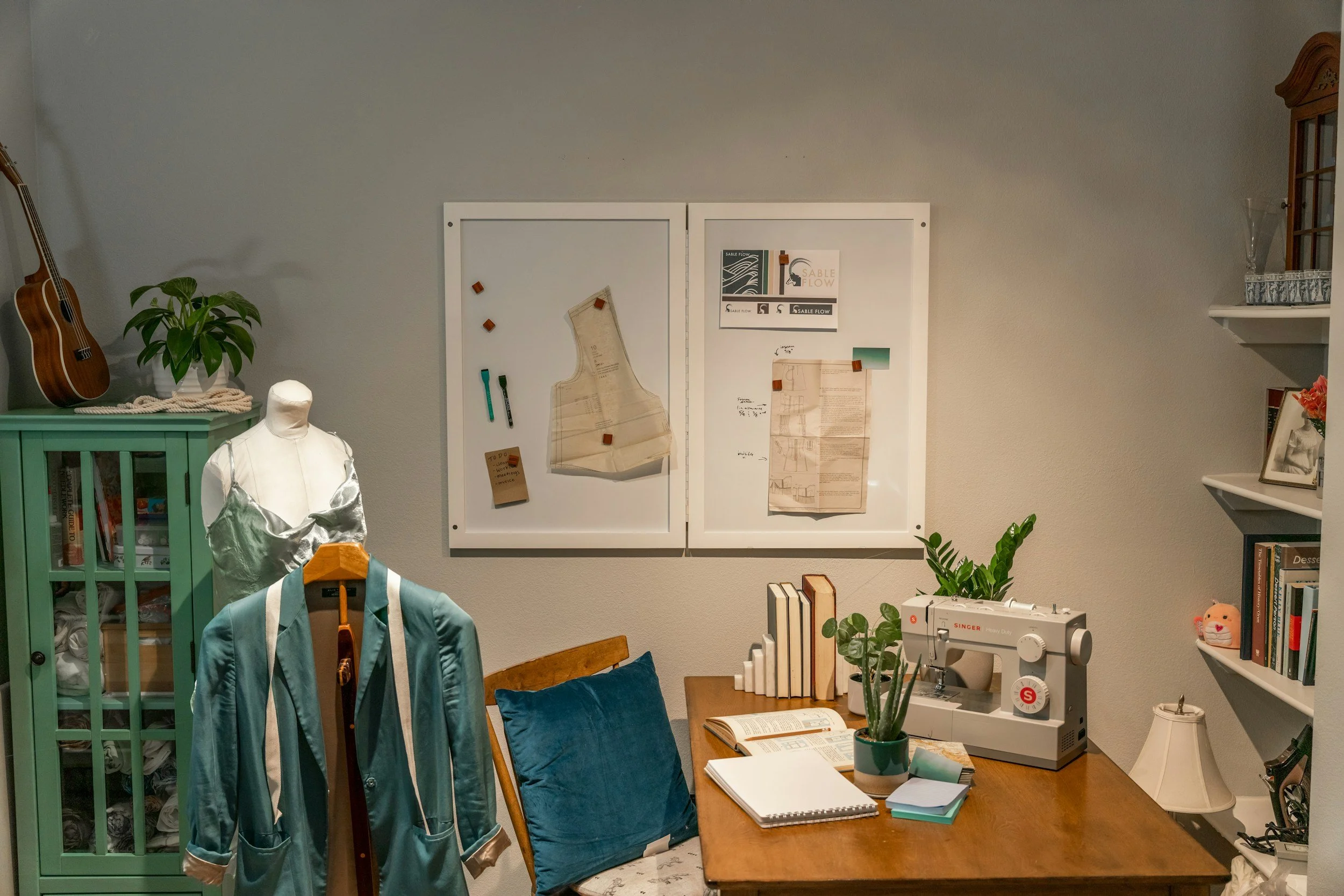 A cozy craft room with a sewing machine on a desk, a mannequin wearing teal and gray clothing, a green cabinet filled with textiles, a photo and notes on a whiteboard, books, plants, and a sewing machine, with a wall shelf with glass vases, and a guitar leaning against the wall.