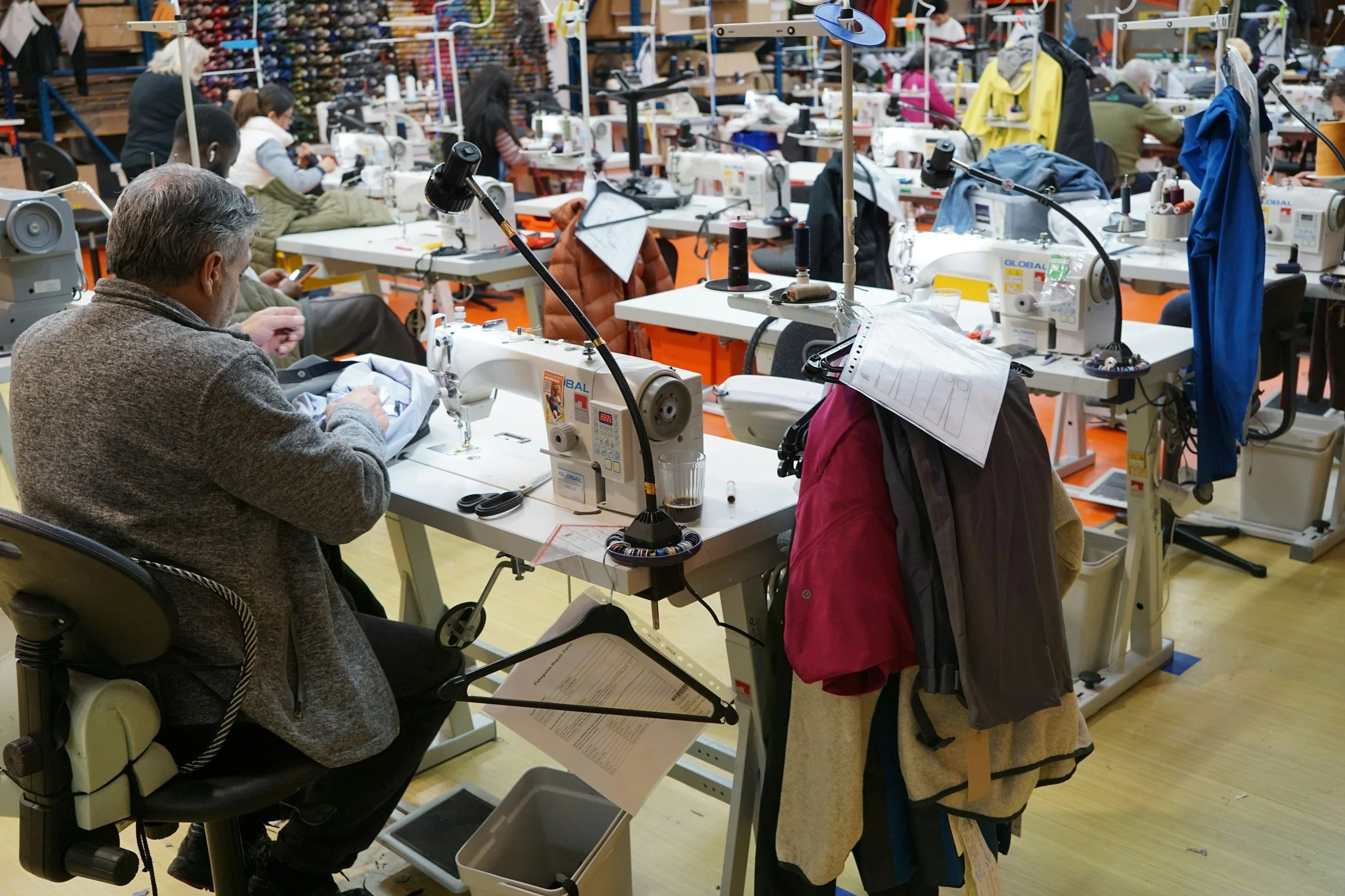 A bustling sewing factory with multiple workers at sewing machines, working on garments. The setting features organized workstations with spools of thread, fabric, and sewing tools, with colorful thread spools lined up on shelves in the background.