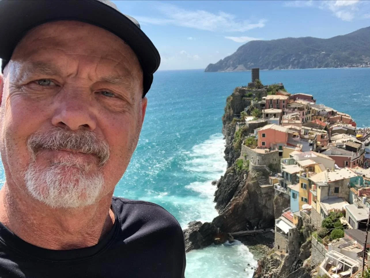 Al stands in the left forefront of the image, wearing a black shirt and black baseball cap. Over his shoulder in as a backdrop of a beautiful Italian coastline.