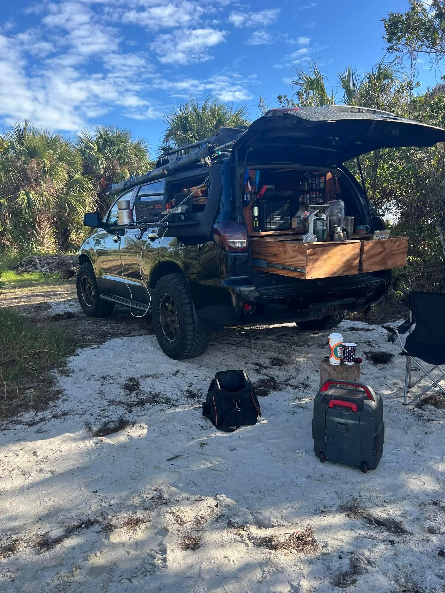 A black Toyota Sequoia is parked in a tropical landscape. The truck, heavily customized, is open to reveal a full camping kitchen setup.