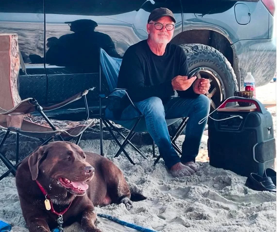In a black shirt, jeans, and a ball cap, Al sits in a folding chair on the beach in front of his black Toyota Sequoia and, on the ground at his feet, his chocolate lab.