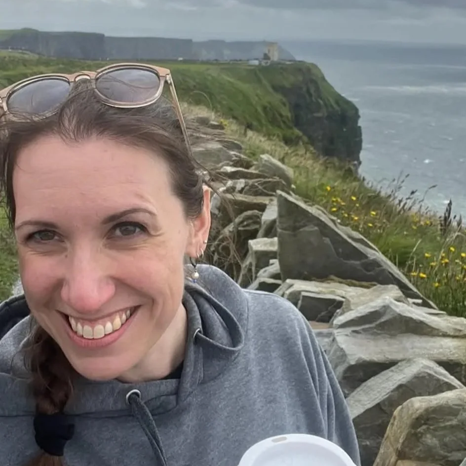 Rachel smiles into the wind, standing in front of the rocky coastline of the Cliffs of Moher in Ireland.