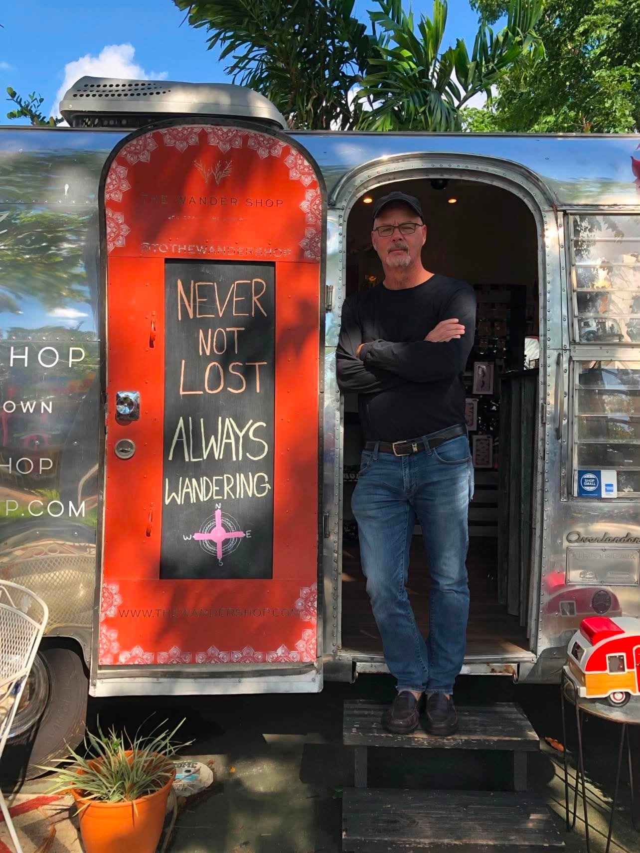 Al stands in the doorway of an Airstream camper, wearing a long-sleeved black shirt and jeans and with arms and legs crossed. The inside of the open and colorfully-painted camper door  reads "Never Not Lost, Always Wandering."